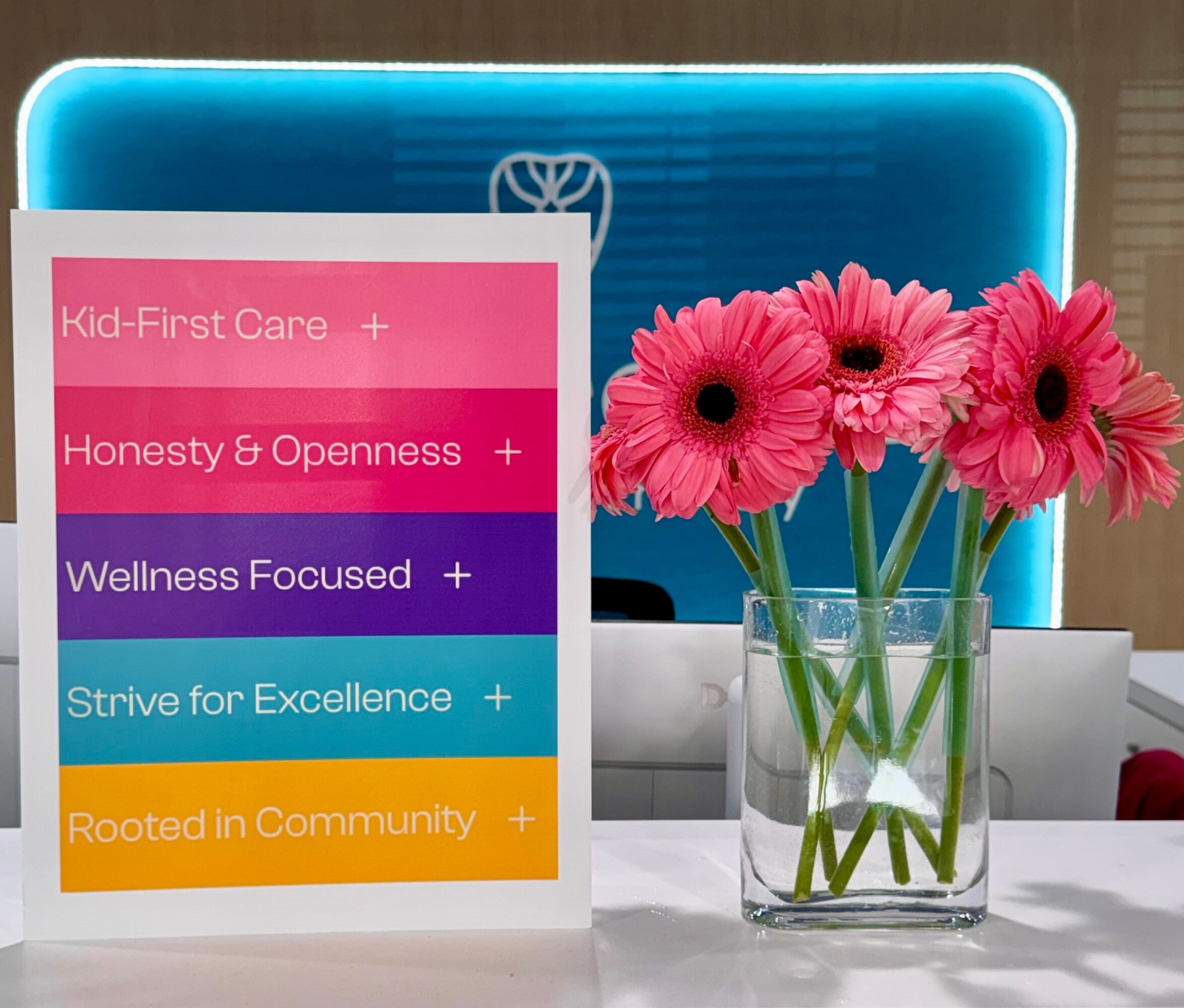 Pink flowers in a glass vase on a table with a colorful sign and a blue laptop in the background.