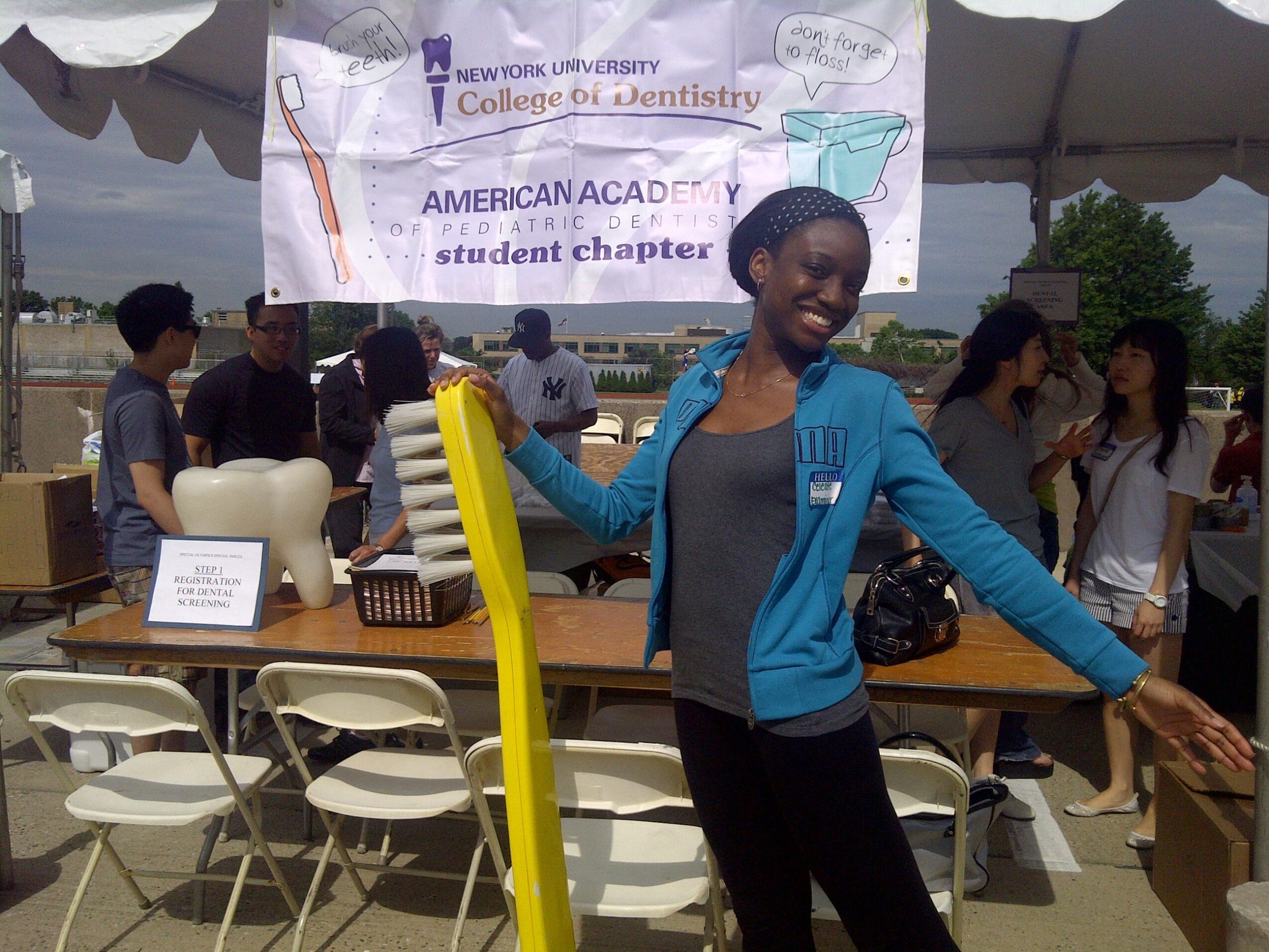 Smiling woman in blue jacket at outdoor event with tables, chairs, and a banner in the background.