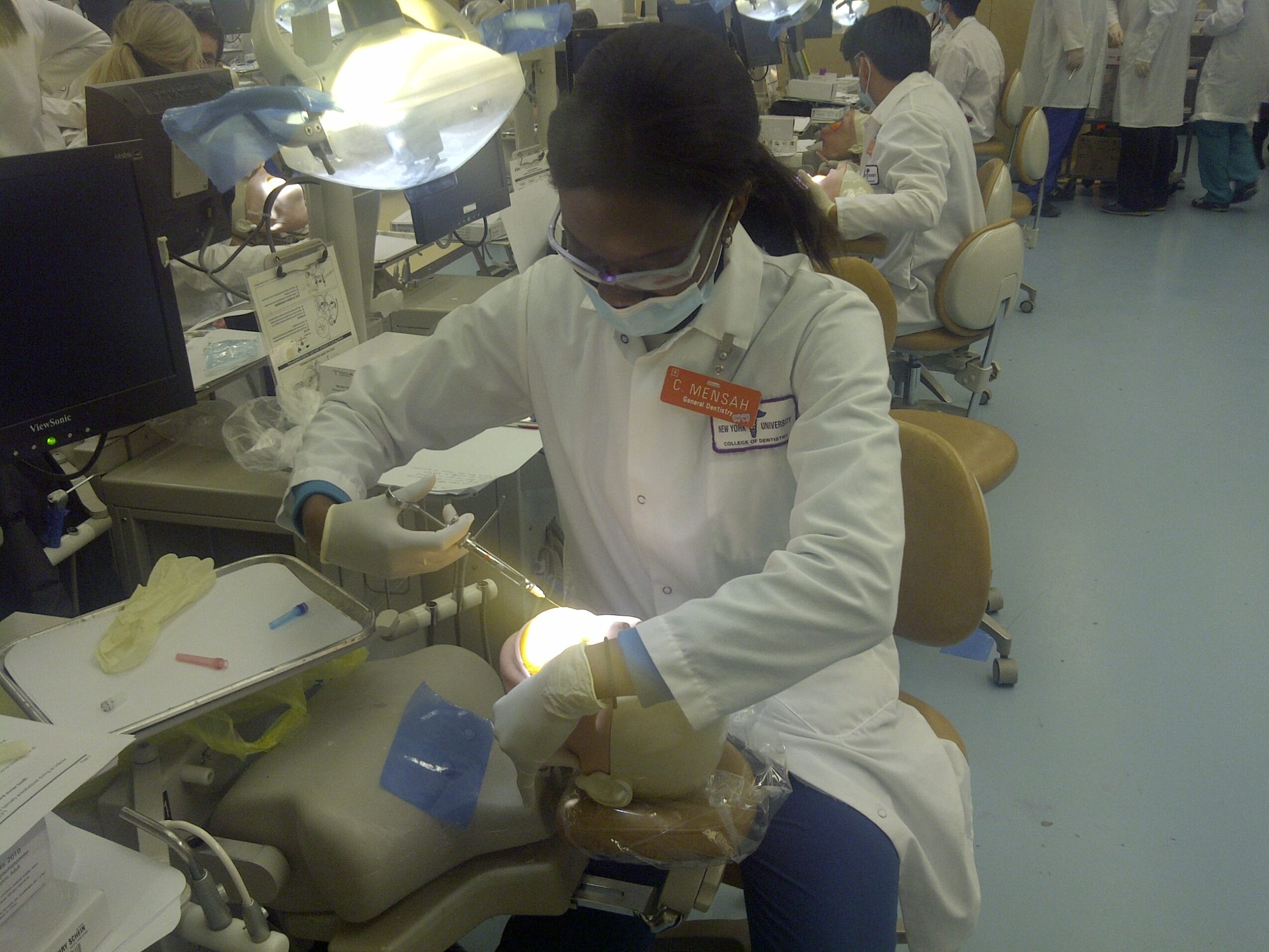 A healthcare professional in a white coat and mask performs a procedure in a busy clinical setting.