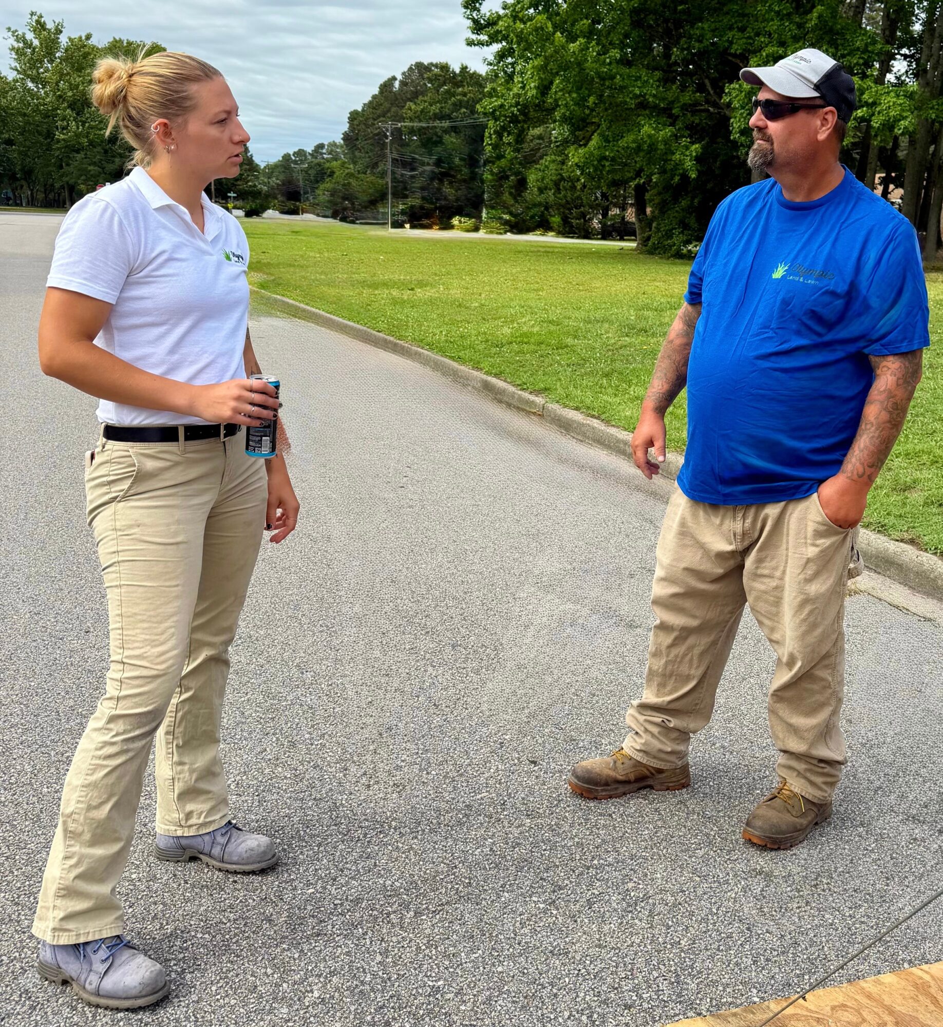 Two people standing outdoors on a paved path, talking, with trees and grass in the background.