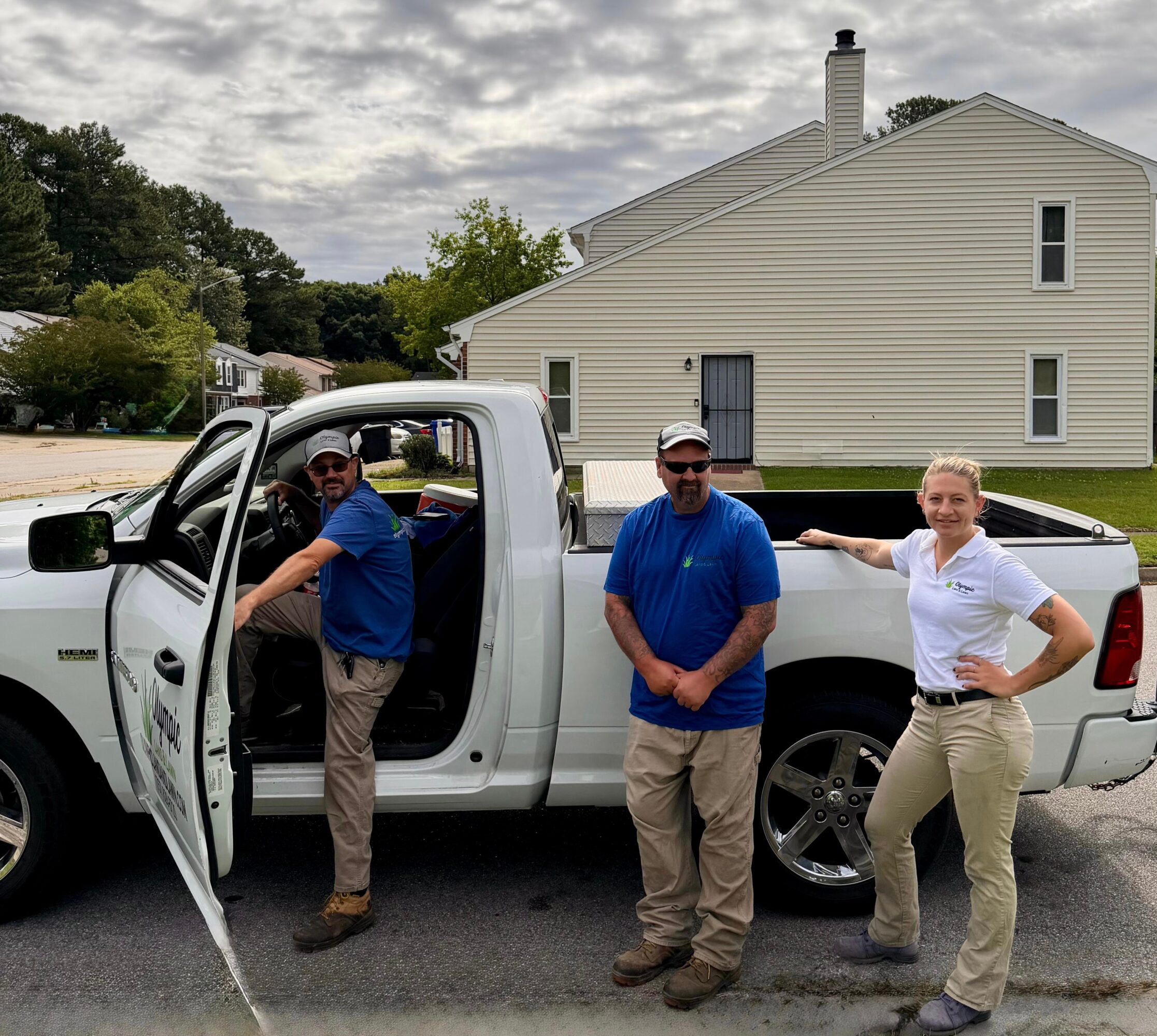 Three people standing next to a white pickup truck on a street, with a house and trees in the background.