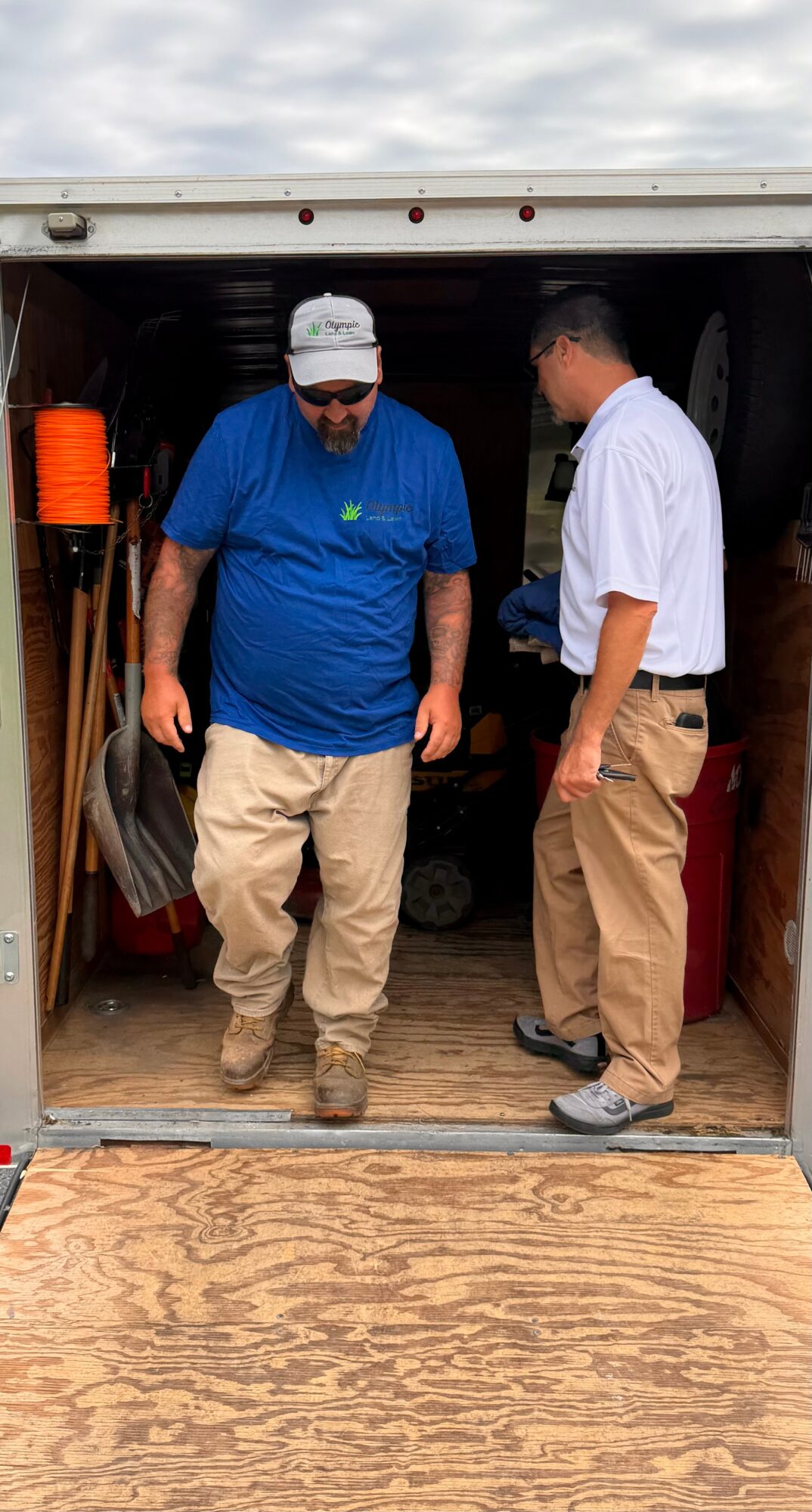 Two men standing in a storage unit, one in a blue shirt and beige pants, the other in a white shirt and beige pants, looking down.