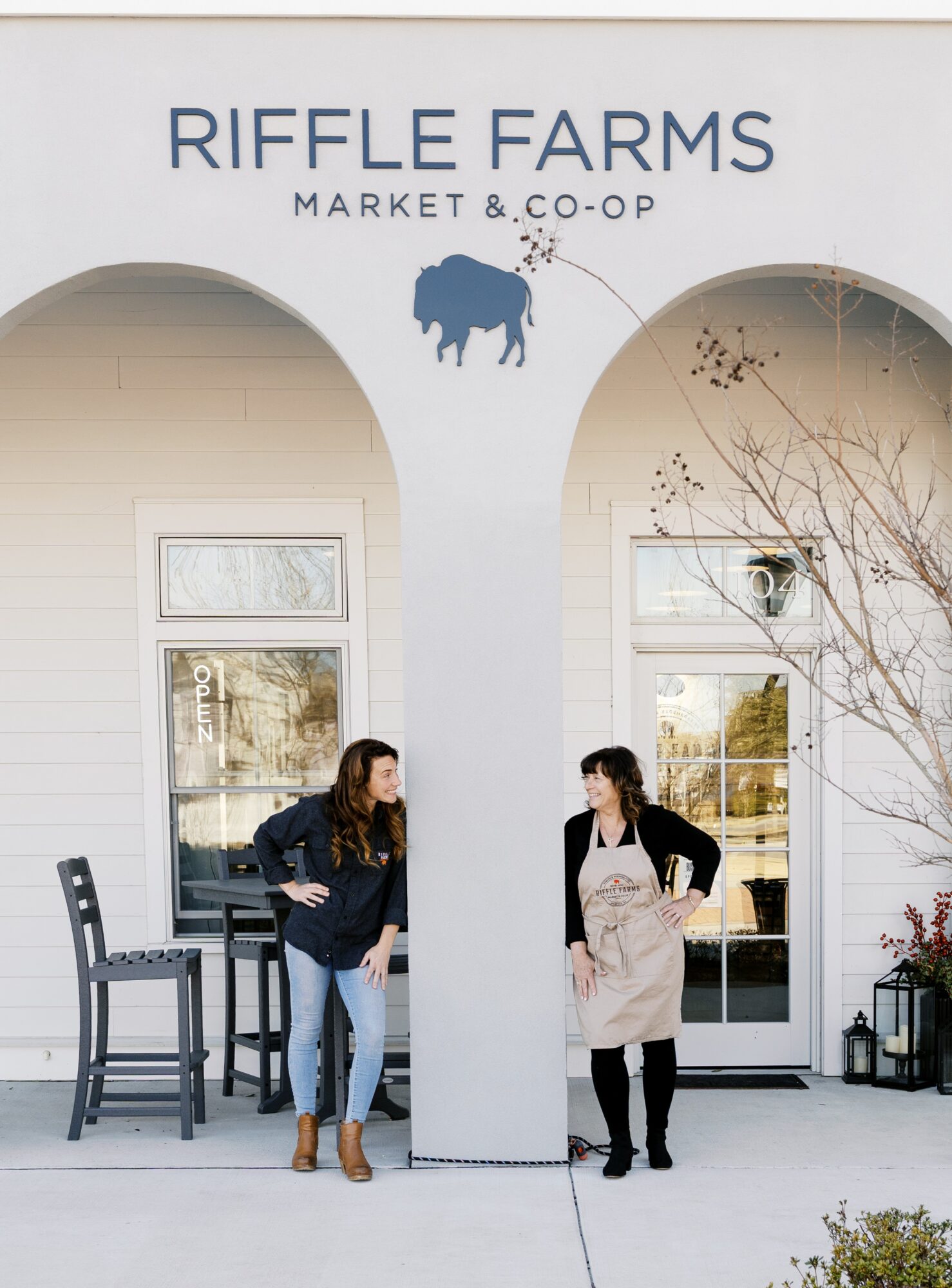 Two women stand outside Riffle Farms Market & Co-Op, talking near a white building with arched entrances and a sign with a buffalo silhouette.