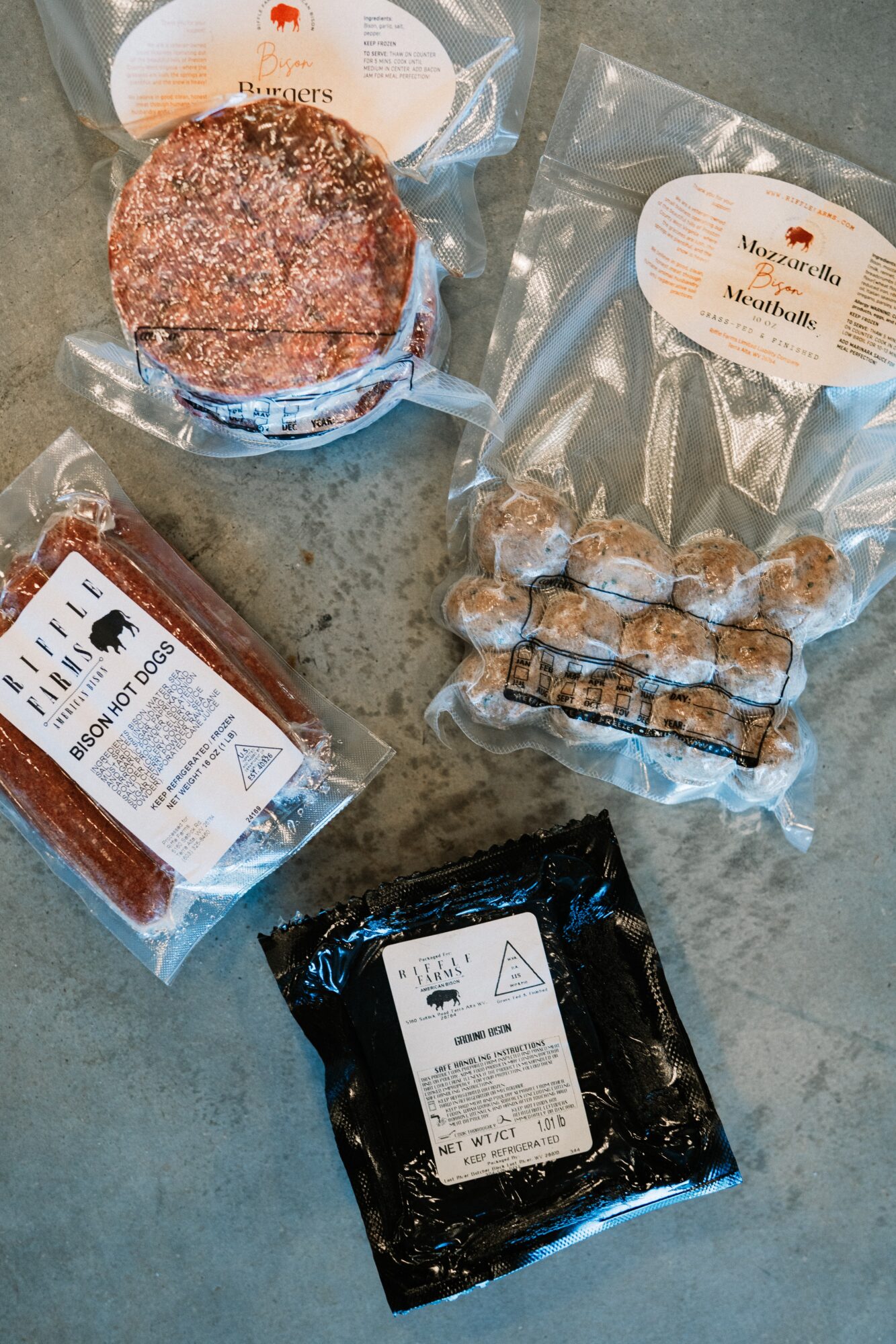 Four packaged food items on a gray surface: a round cake, a bag of small round snacks, a rectangular block, and a black package with a white label.