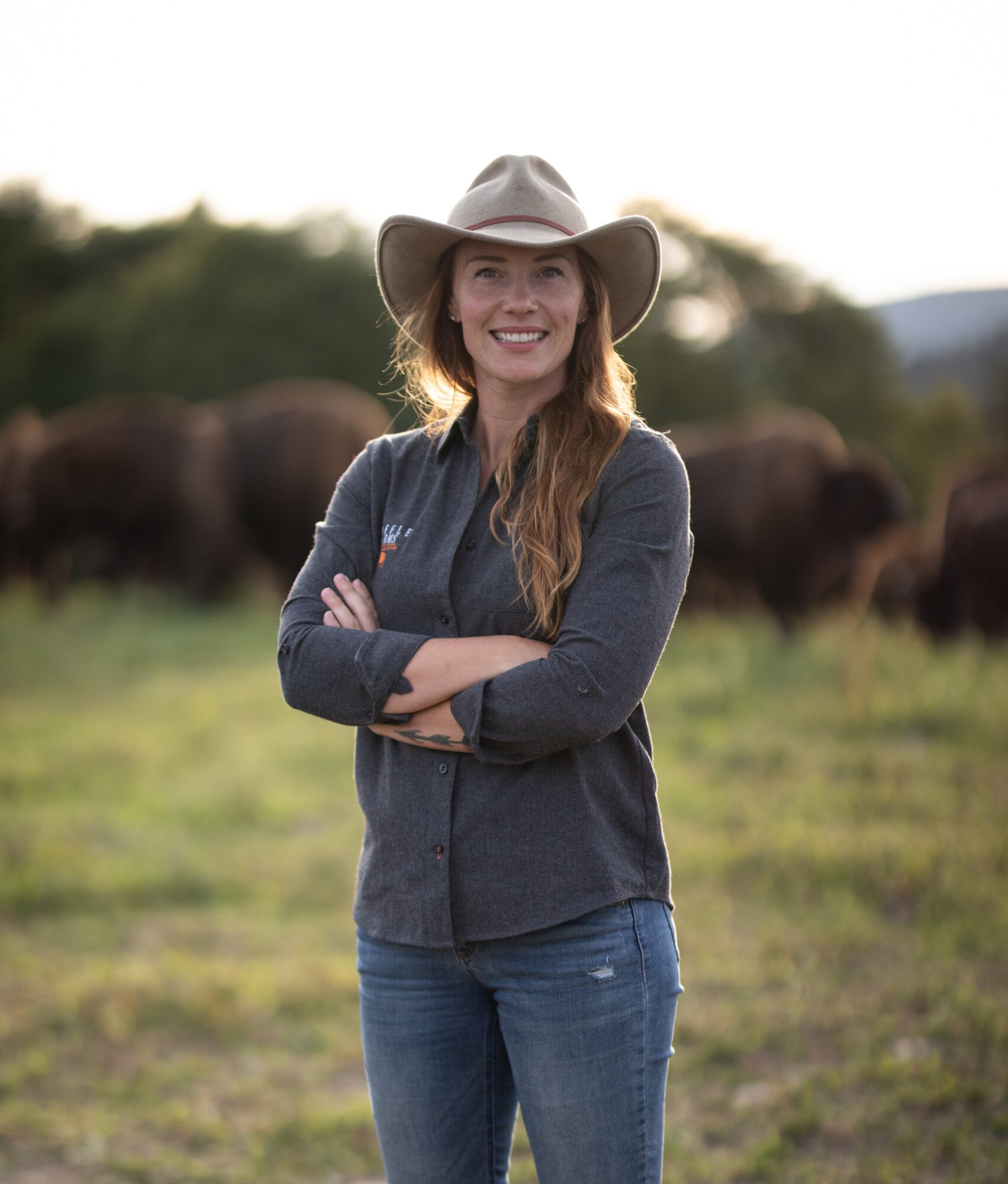 Woman in cowboy hat smiling with arms crossed outdoors, bison in background, grassy field, natural landscape, daytime.