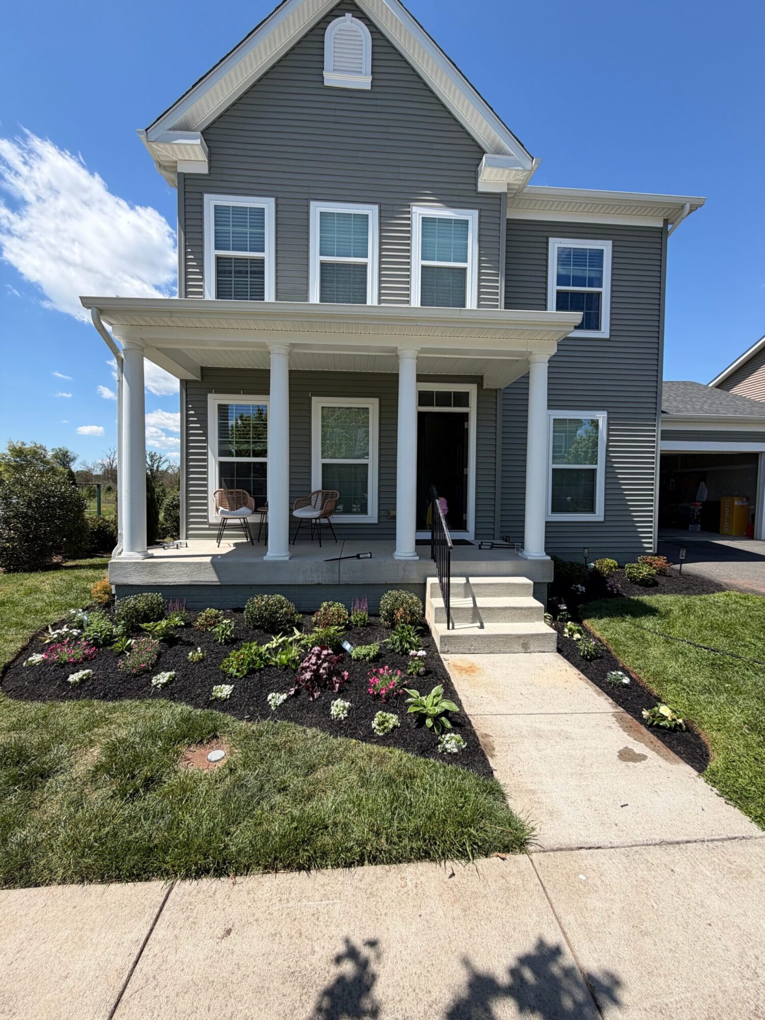 Front view of a gray house with a porch, garden bed, and sidewalk, under a blue sky with clouds.