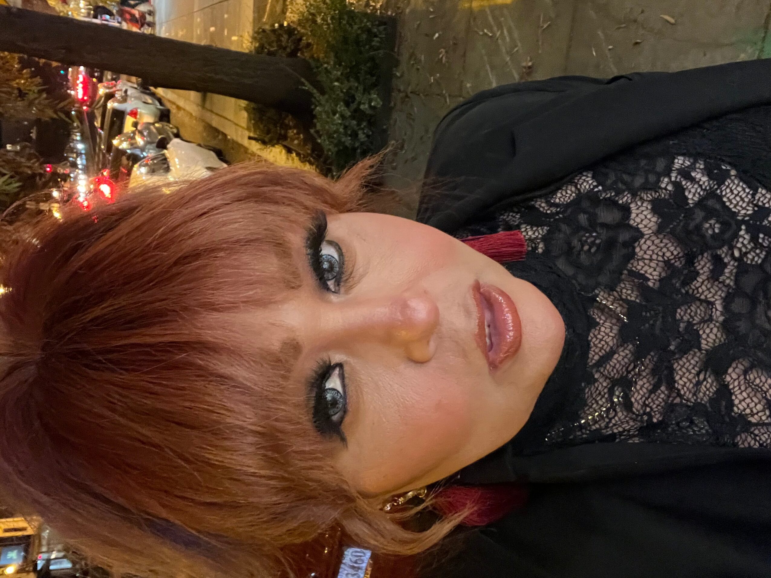 Woman with red hair and blue eyes wearing black lace top, outdoors at night with city lights in background.