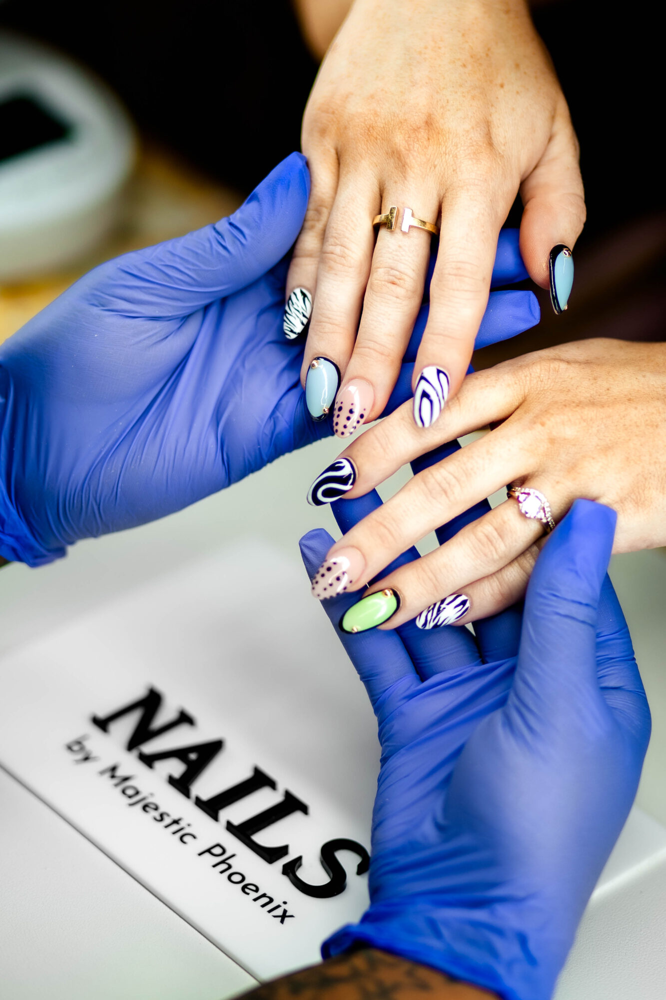 Person receiving a manicure, hands with decorated nails, gloves worn by technician, nail salon setting, NAILS sign visible.