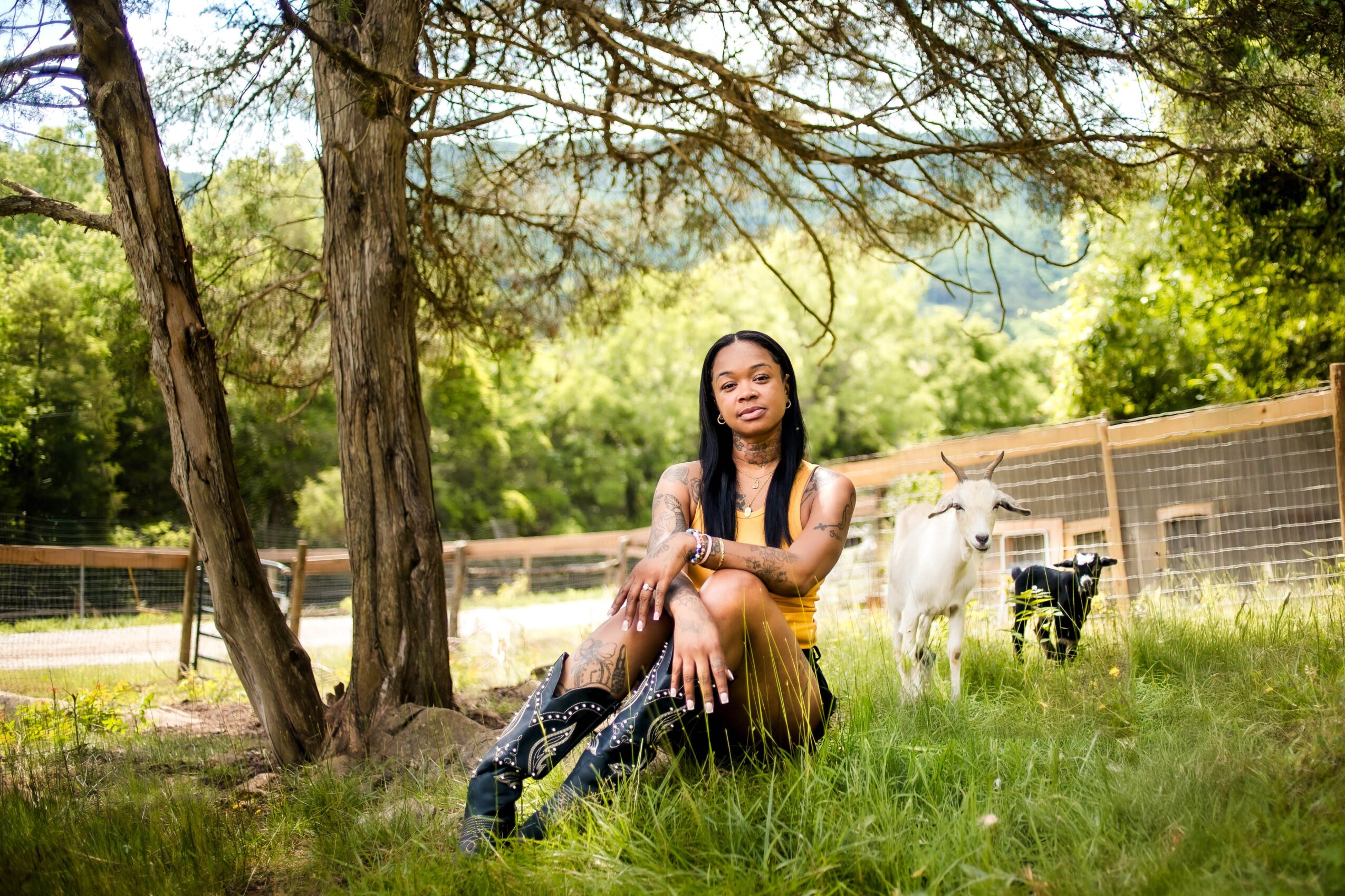 Young woman sitting on grass near trees with goats in background, outdoors on sunny day.