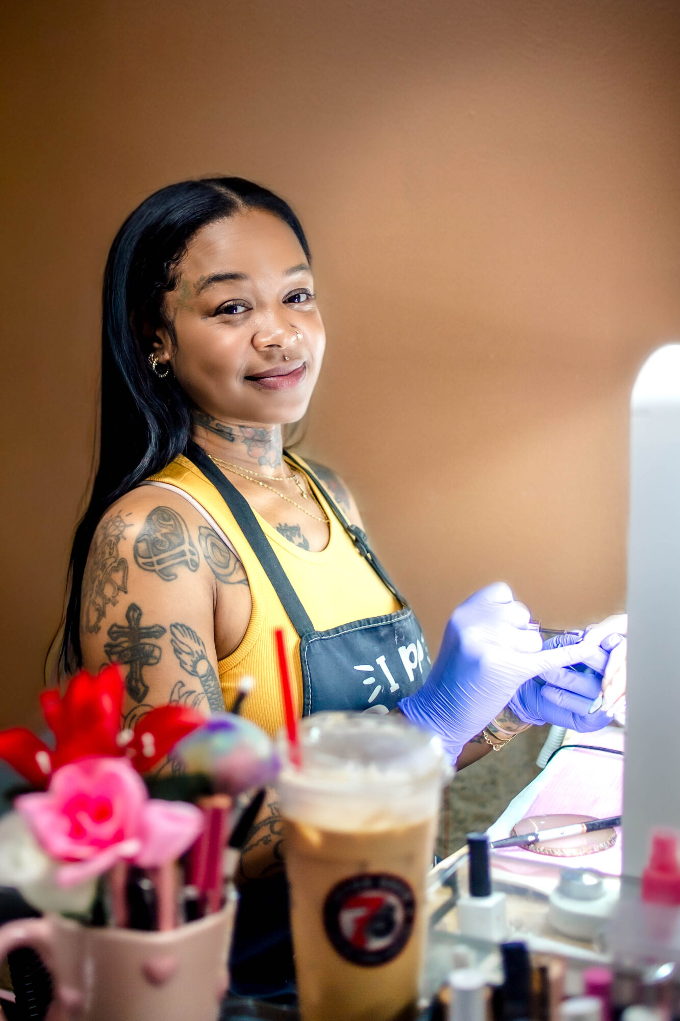 Young woman with tattoos smiling, wearing a yellow top and apron, working with tools at a nail salon, with pink flowers and drinks in foreground.