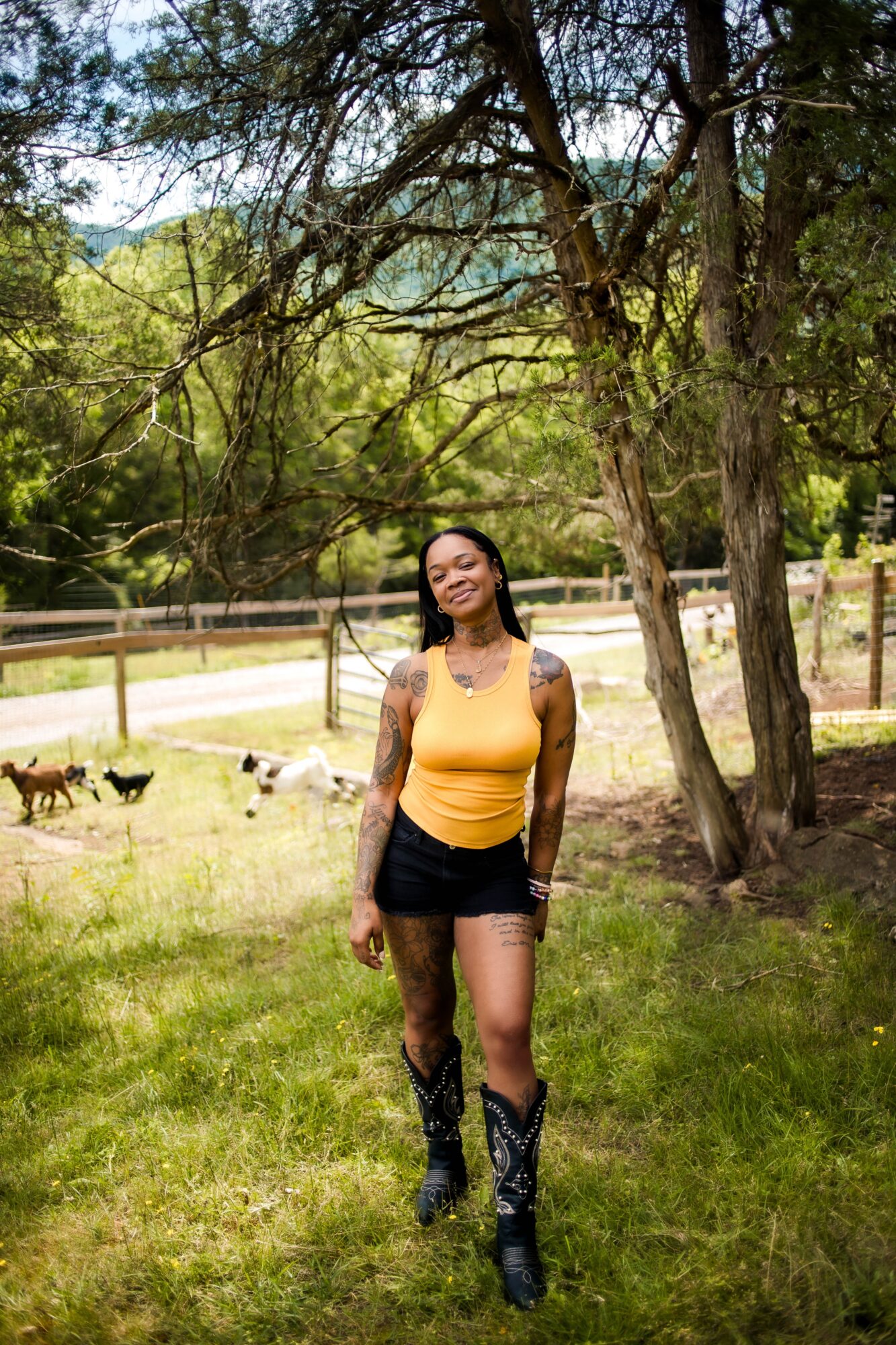 Woman standing outdoors in a grassy area with trees, wearing a yellow top and black shorts, smiling.