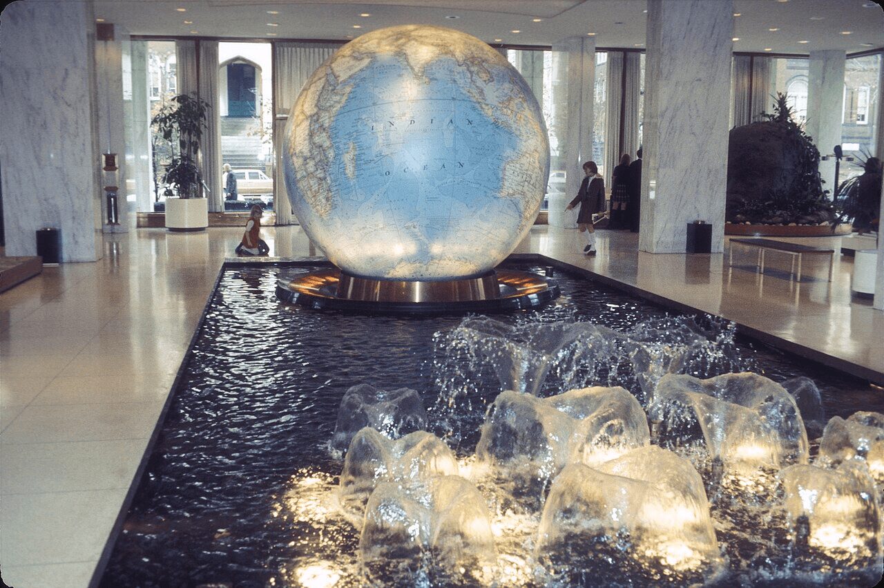 A globe sculpture sits above a water fountain with bubbling rocks inside a modern building lobby.
