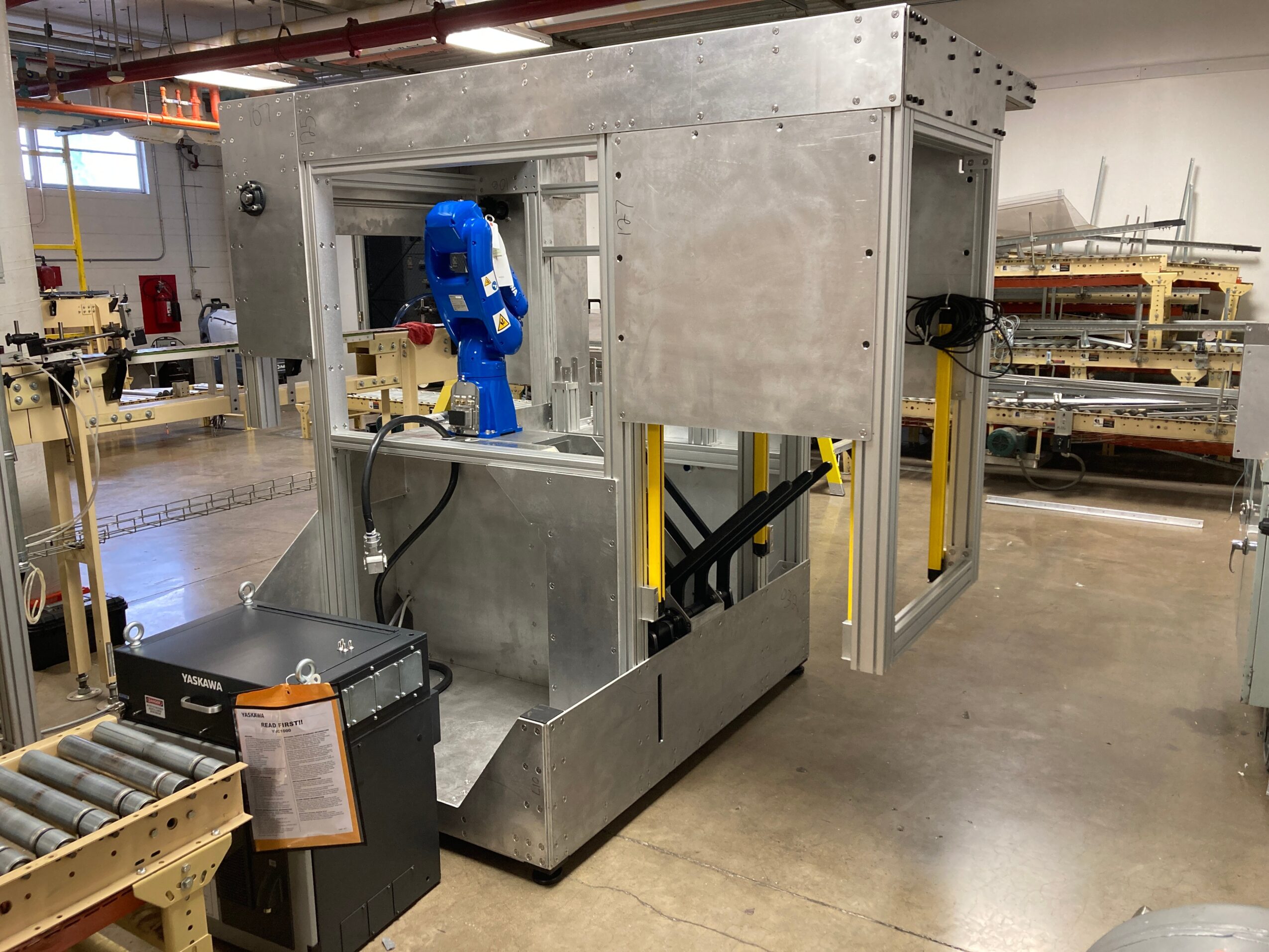 Large metallic machine with blue robotic arm inside a workshop, surrounded by workbenches and equipment.
