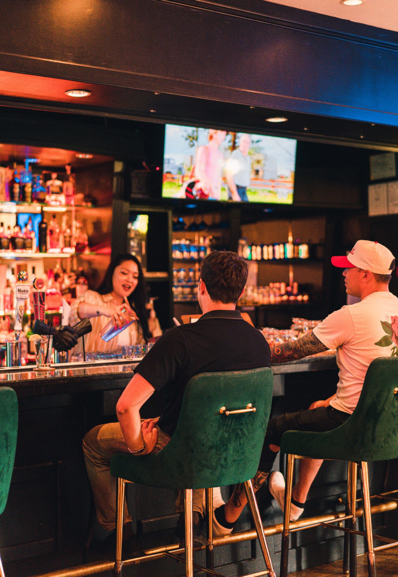 Three people sit at a bar counter with a bartender serving drinks, in a dimly lit setting with shelves of bottles behind.
