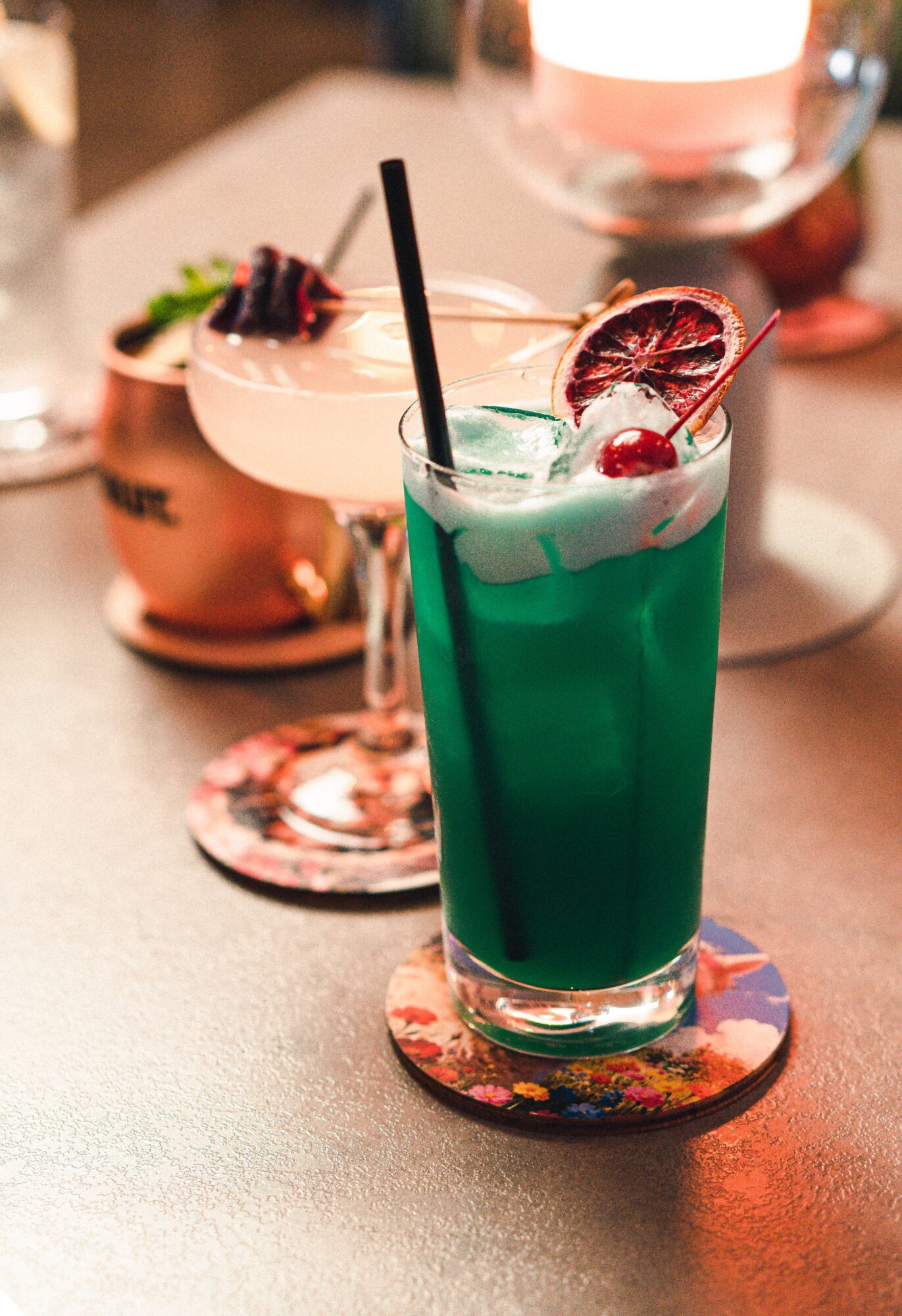 Tall glass with blue-green drink, lime slice, and straw on a colorful coaster, with other drinks in the background.