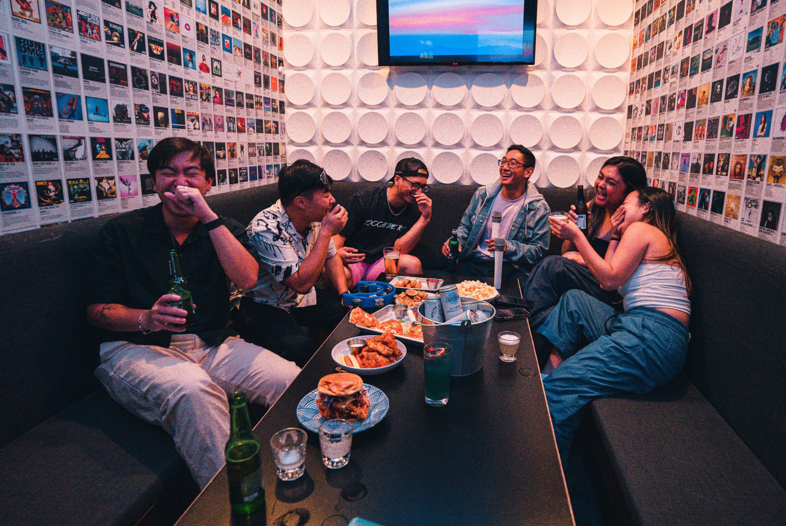 Group of six people sitting around a table with food and drinks, in a room with photo walls and a TV screen.