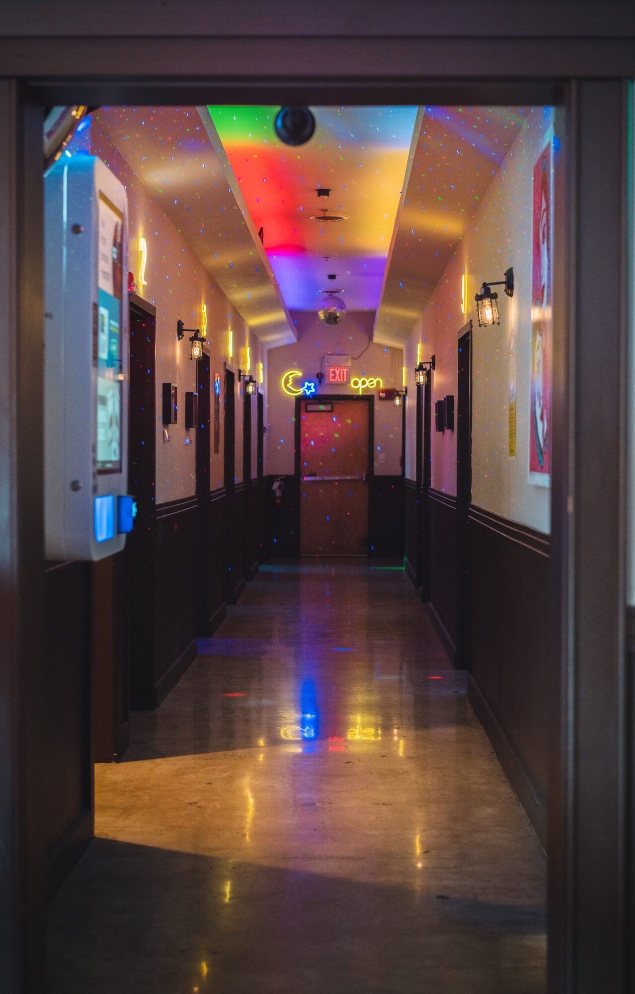 Colorful hallway with rainbow ceiling lights, neon signs, and wall-mounted lights, leading to a door at the end.