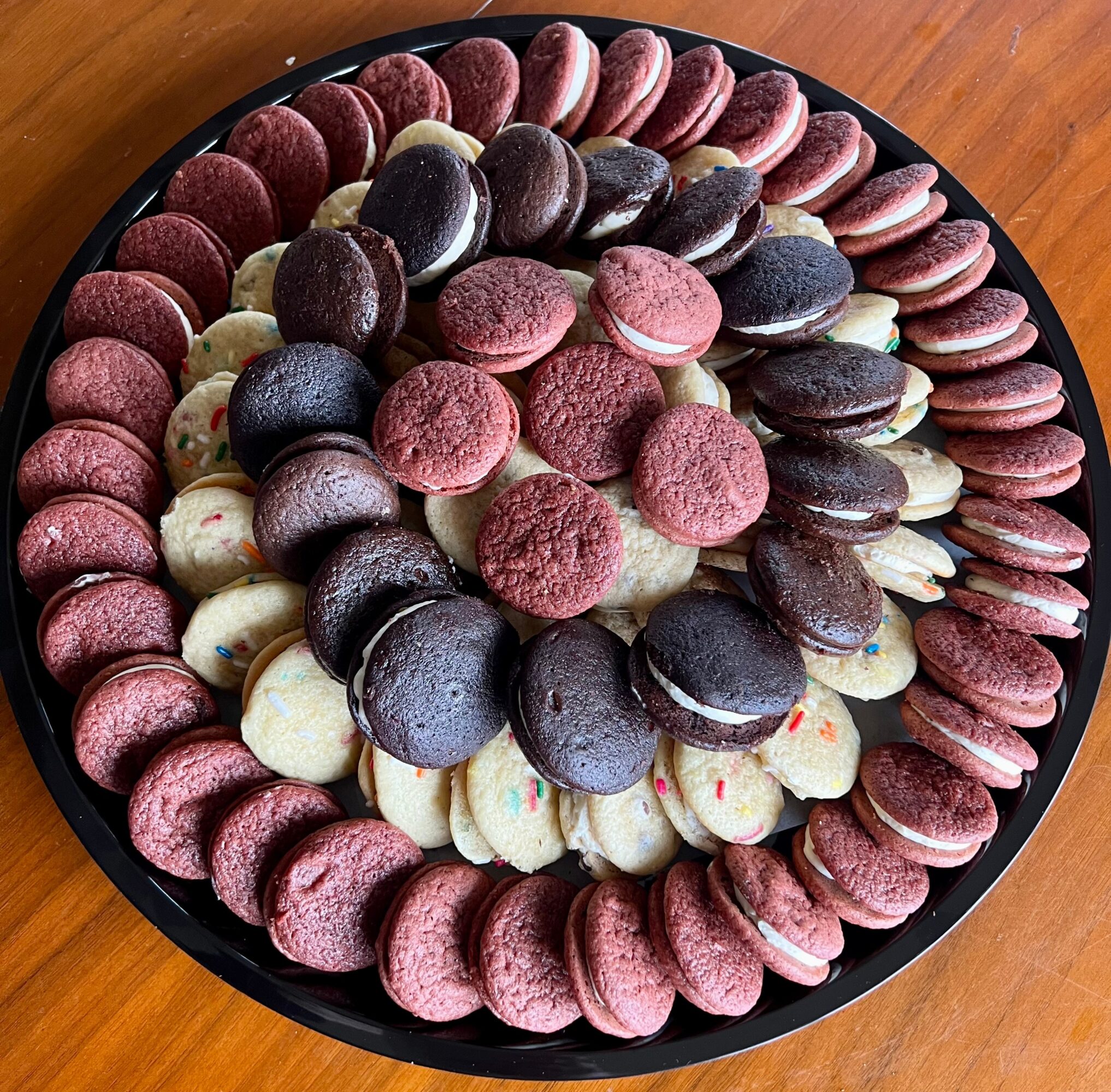 Assorted macarons arranged in a circular pattern on a black tray, with pink, purple, and white colors, on a wooden surface.
