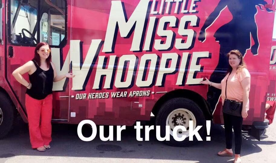 Two women stand in front of a pink bus with a large logo and text, holding a leash attached to a dog. The bus reads 'Little Miss Whoopie' and 'Our heroes wear aprons.'