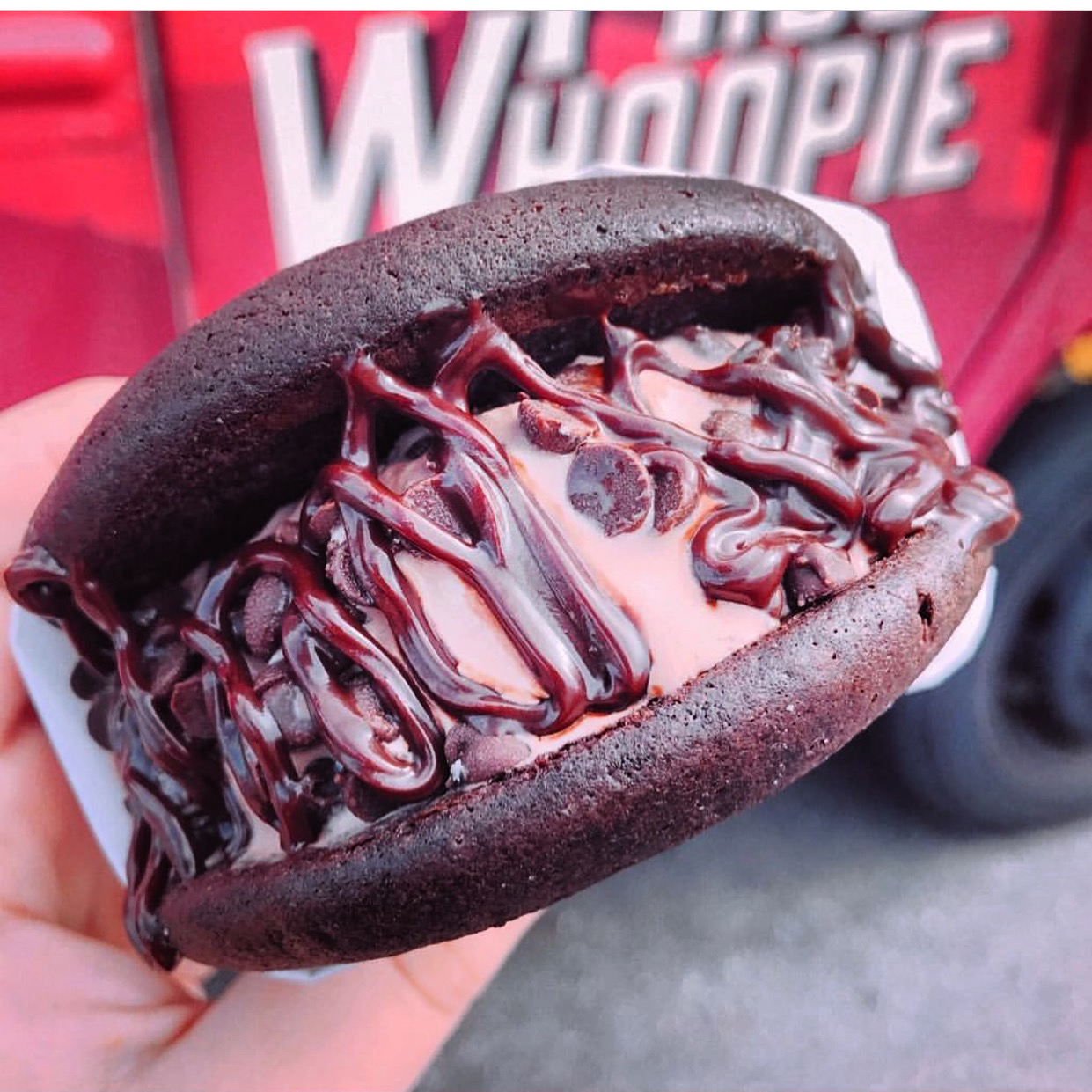 Chocolate ice cream sandwich with chocolate sauce in a black bun, held outdoors with a red vehicle in the background.