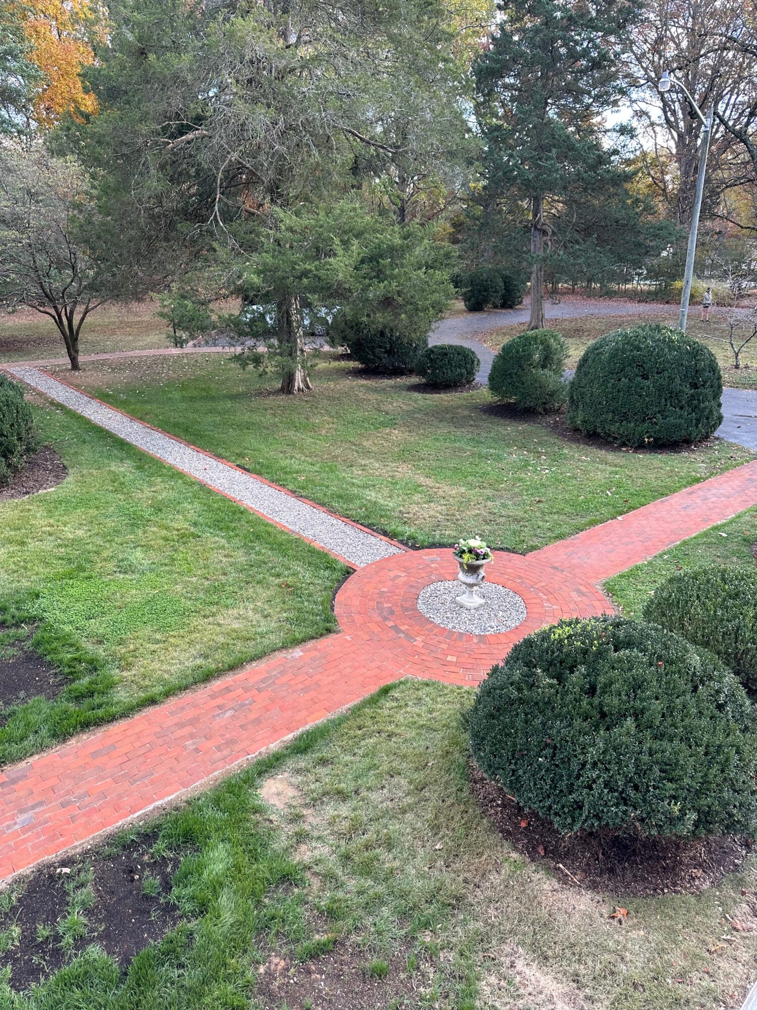 A park with intersecting brick pathways, green grass, trees, and neatly trimmed bushes, viewed from above.
