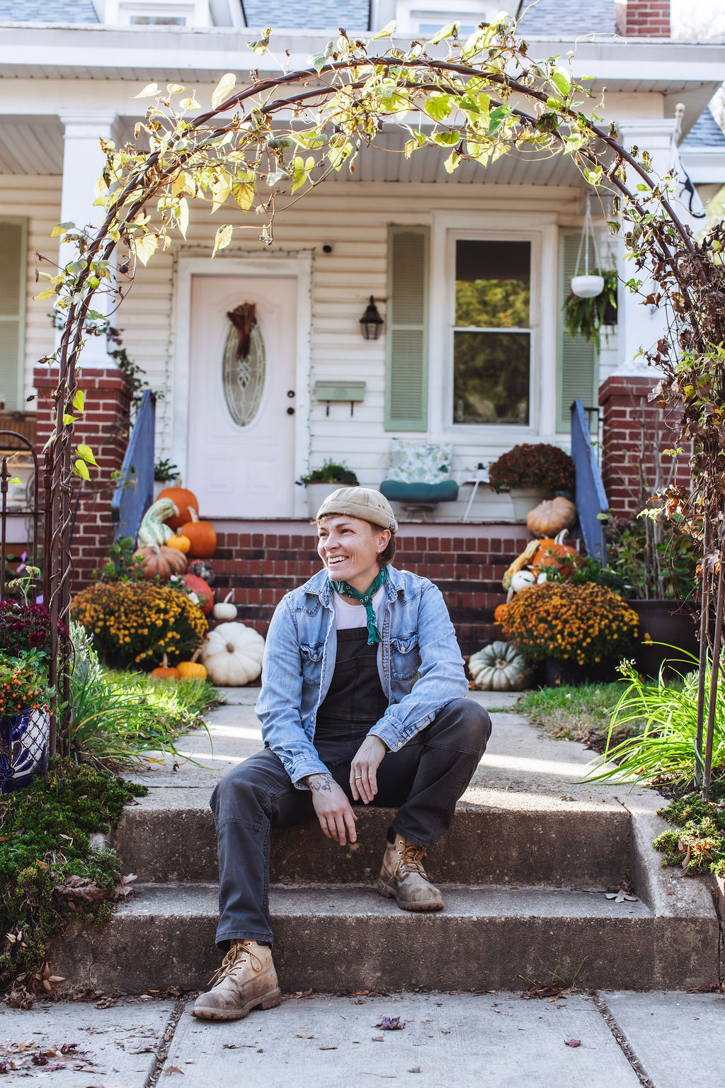 Young man sitting on front steps of a house decorated with pumpkins and fall plants, smiling outdoors.