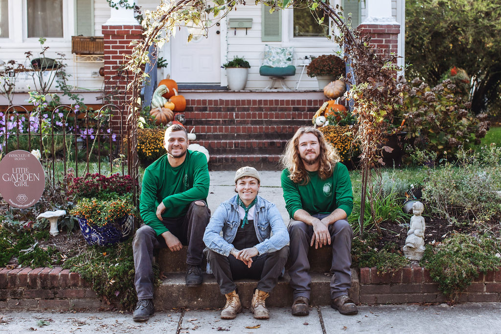 Three people sitting on a brick ledge in front of a house with autumn decorations, smiling outdoors.