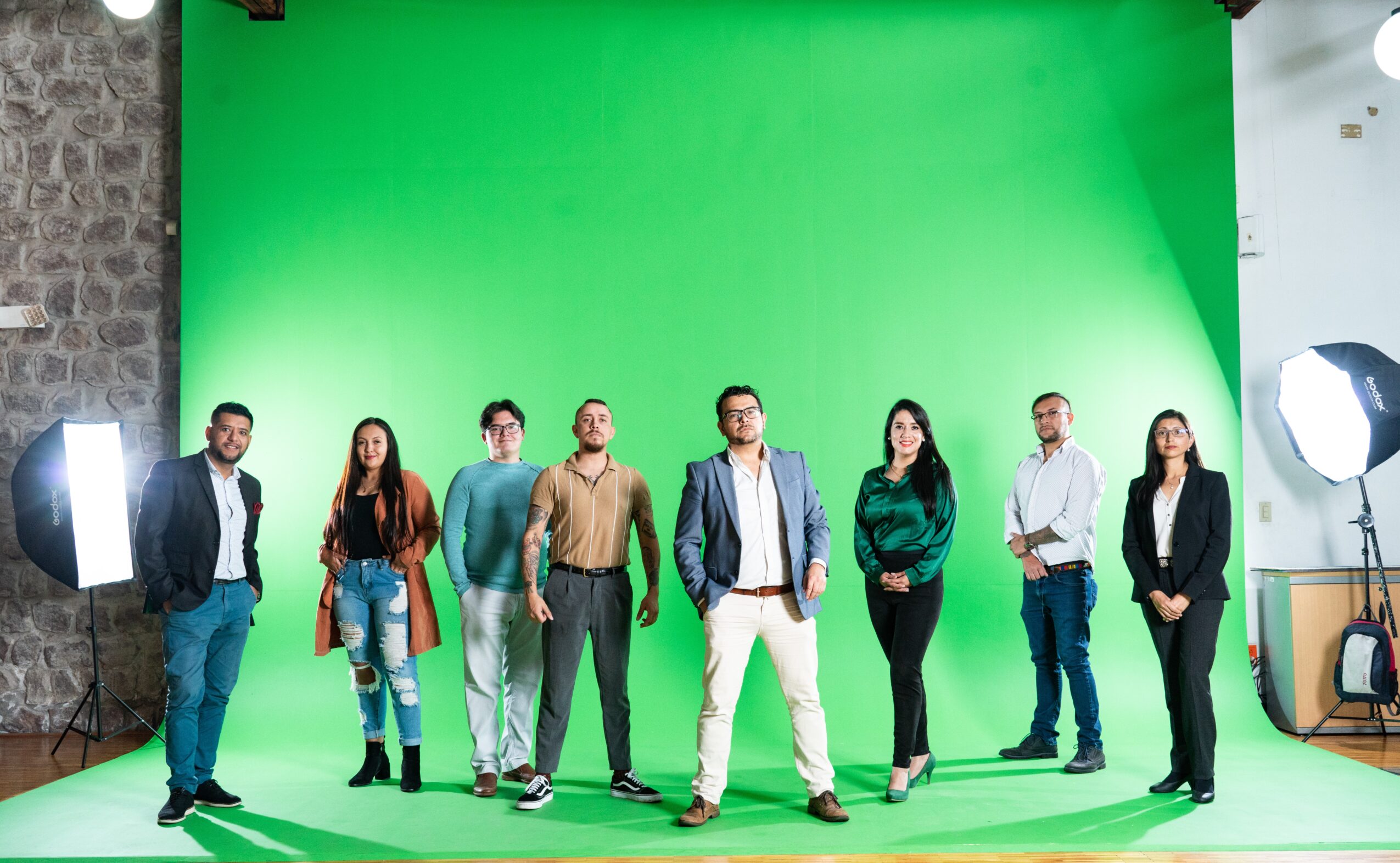 Group of eight people standing in front of a green screen, studio lighting visible, brick wall on the left.