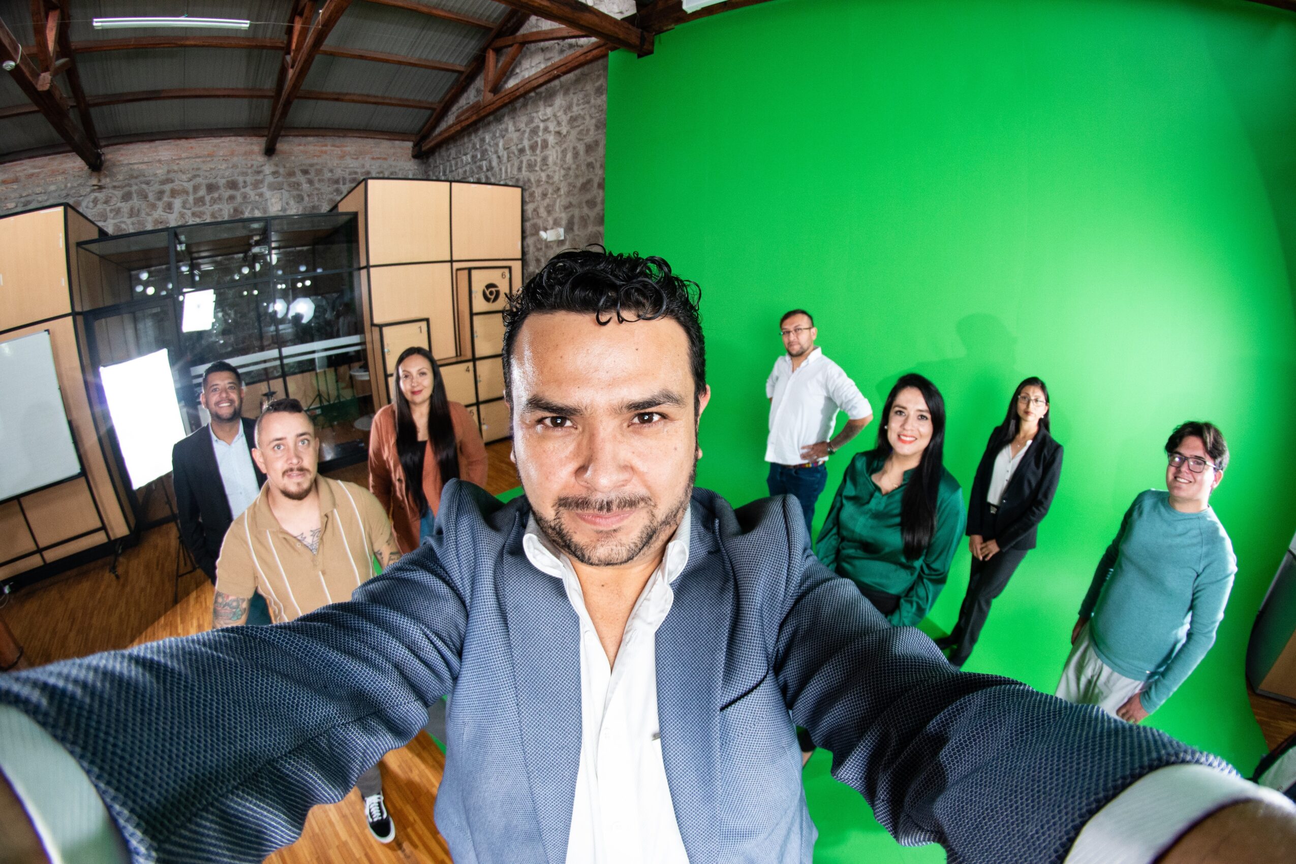 Man taking a selfie in a studio with a green screen and six people in the background.