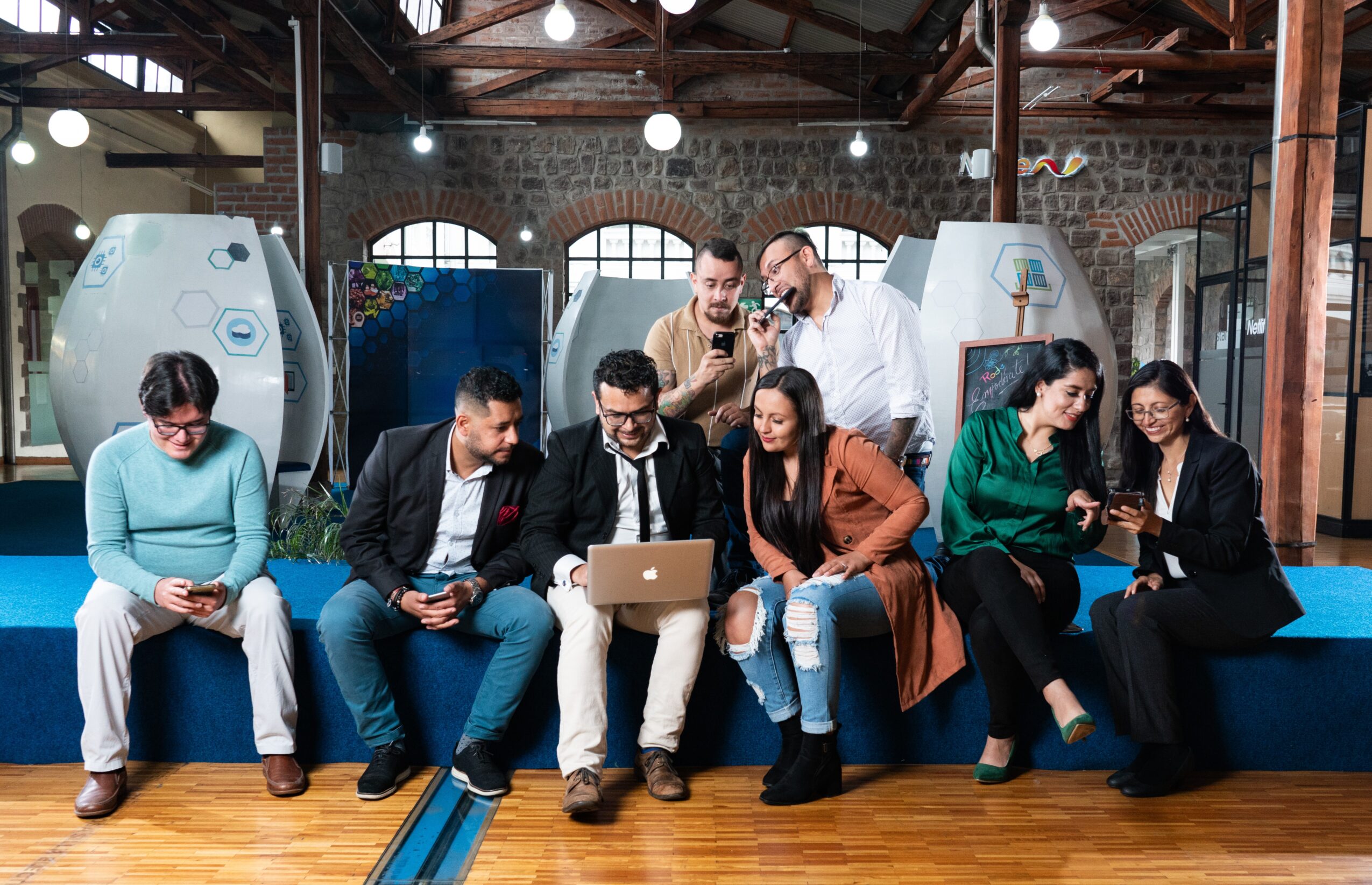 Group of eight people sitting and standing on a blue bench in a modern, industrial-style room with large windows and exposed beams, engaging with electronic devices.