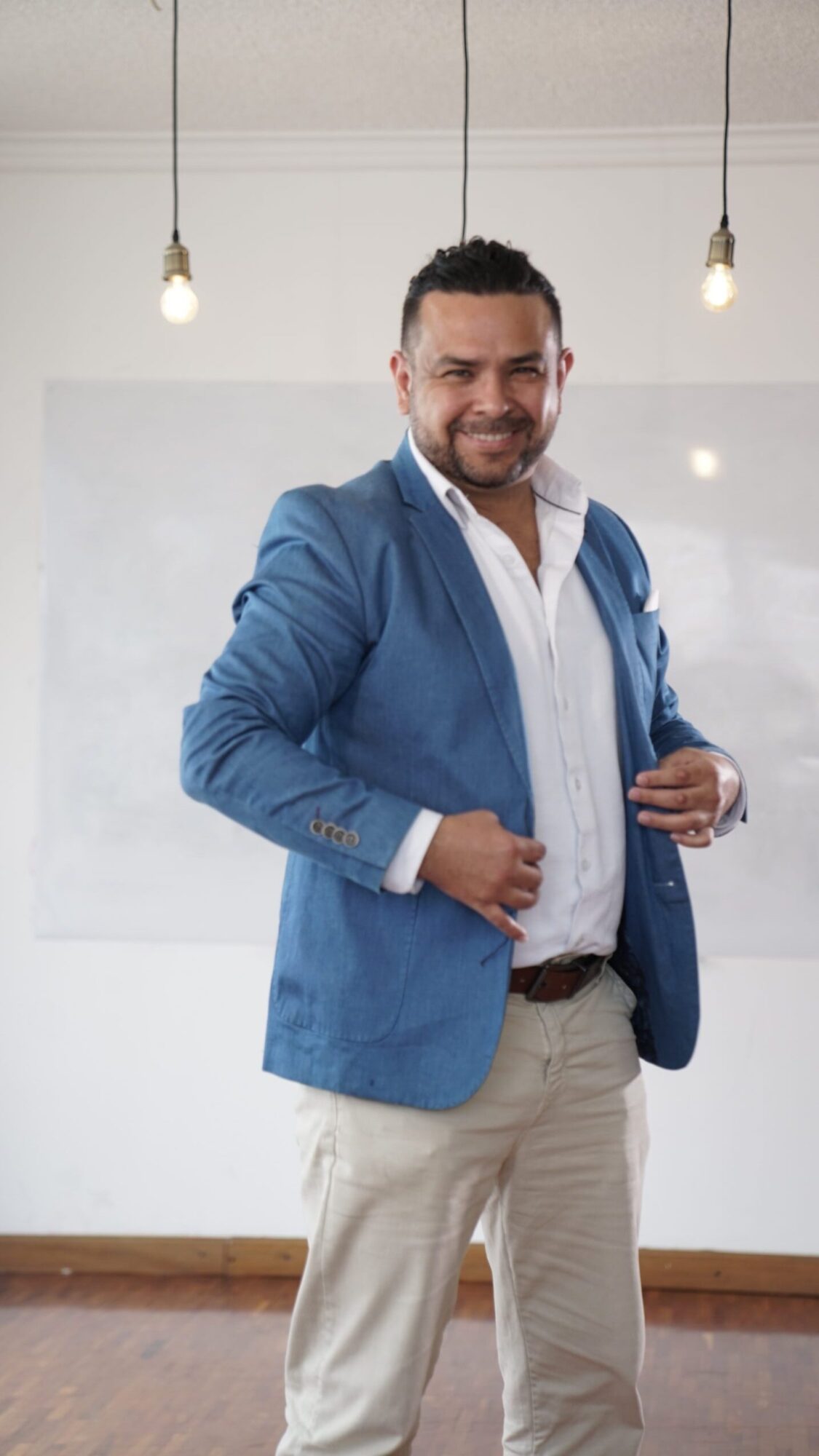 Man in blue blazer and beige pants standing in a room with white walls and wooden floor, smiling and holding a microphone.