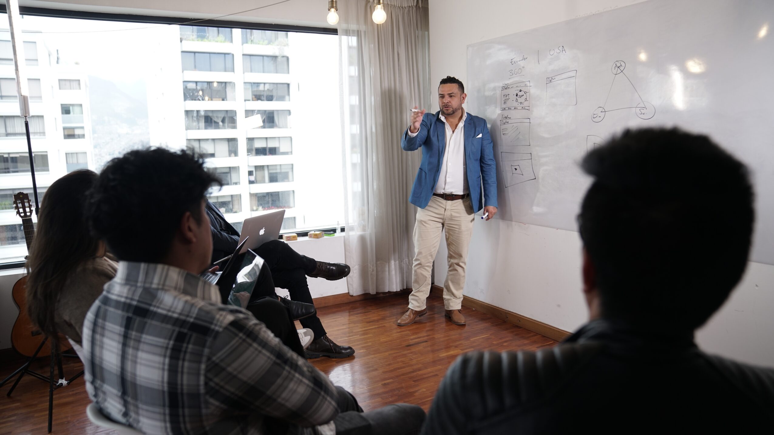 Man in blue jacket giving a presentation to a group in a room with large windows and a whiteboard.