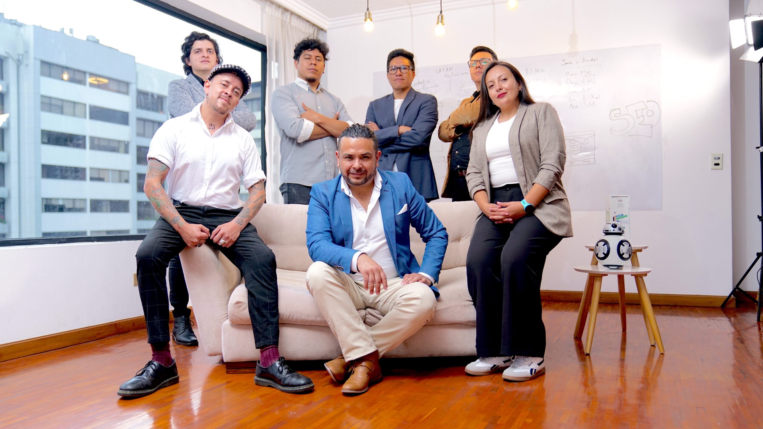 Group of six diverse professionals in a modern office, some standing and some sitting, with large windows and city view