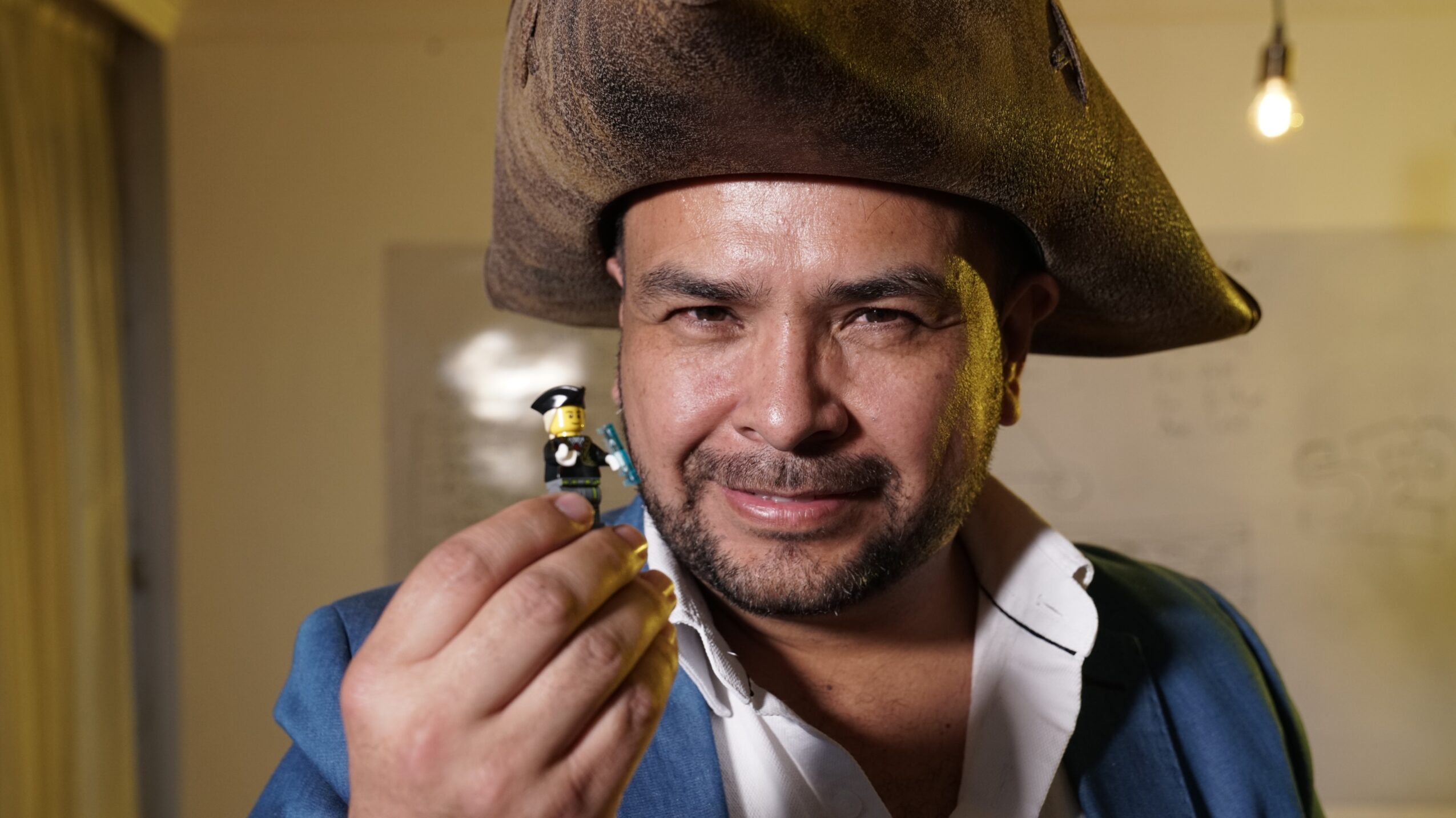 Man dressed as a pirate holding a small toy figure, smiling, in a room with warm lighting.