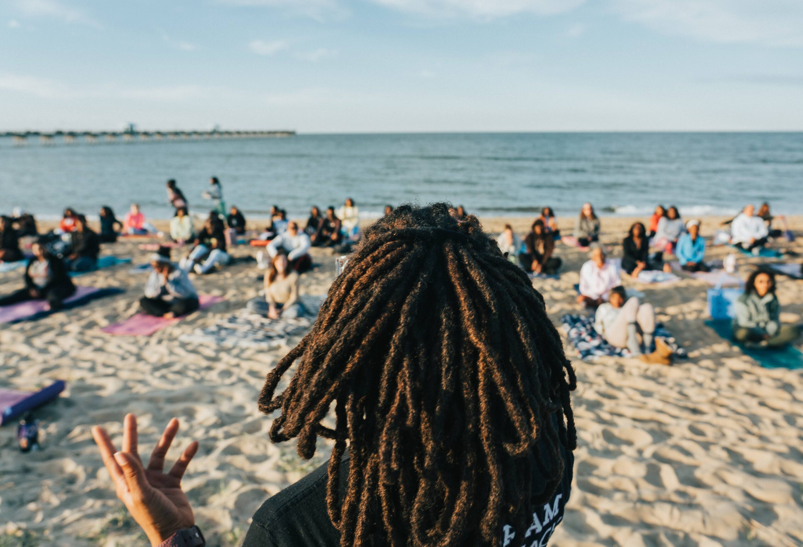 Person with dreadlocks facing a crowded beach with people sitting on sand and near the water, ocean in background