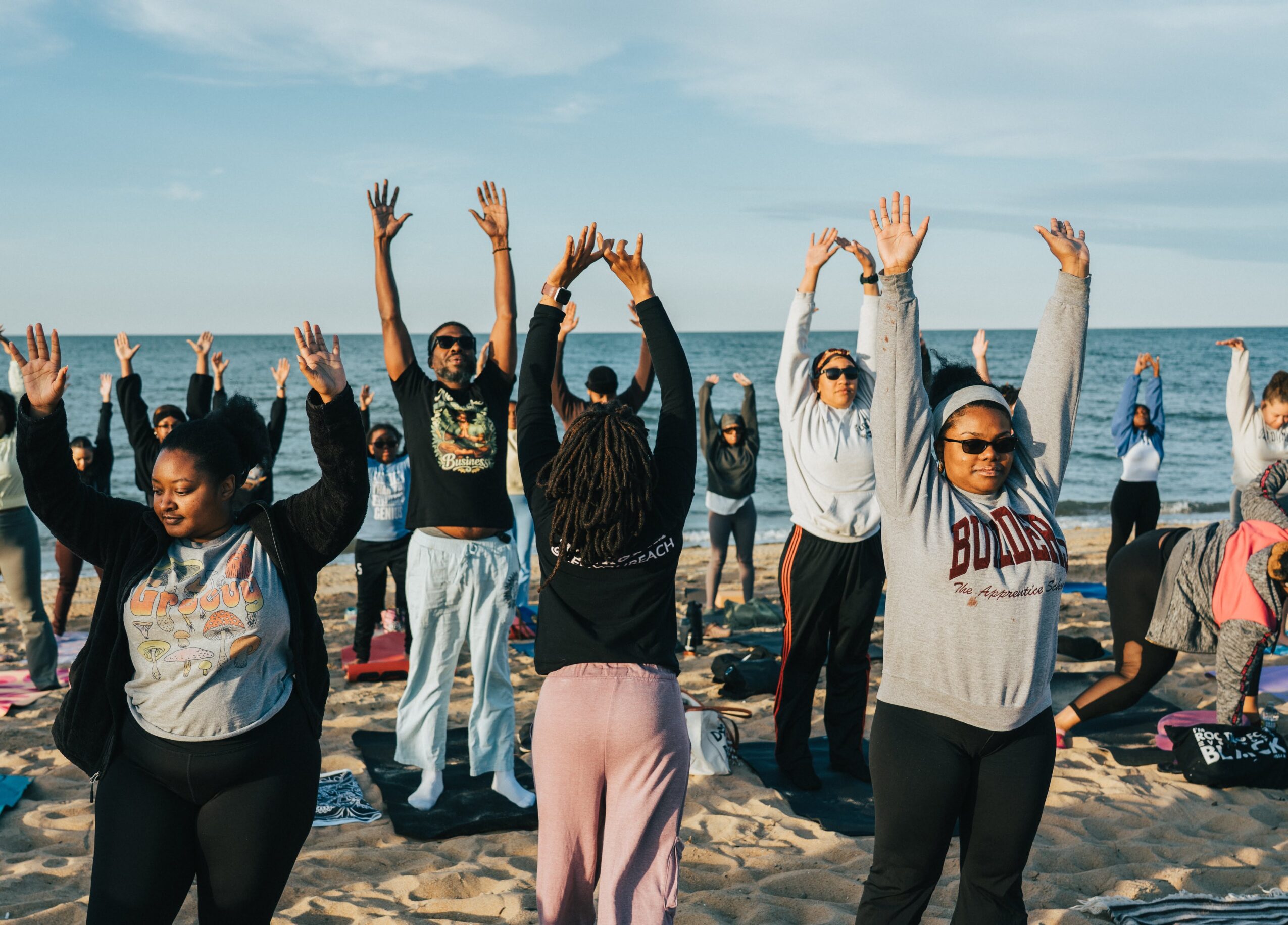 Group of people practicing yoga on a beach, raising arms, with ocean and sky in background.
