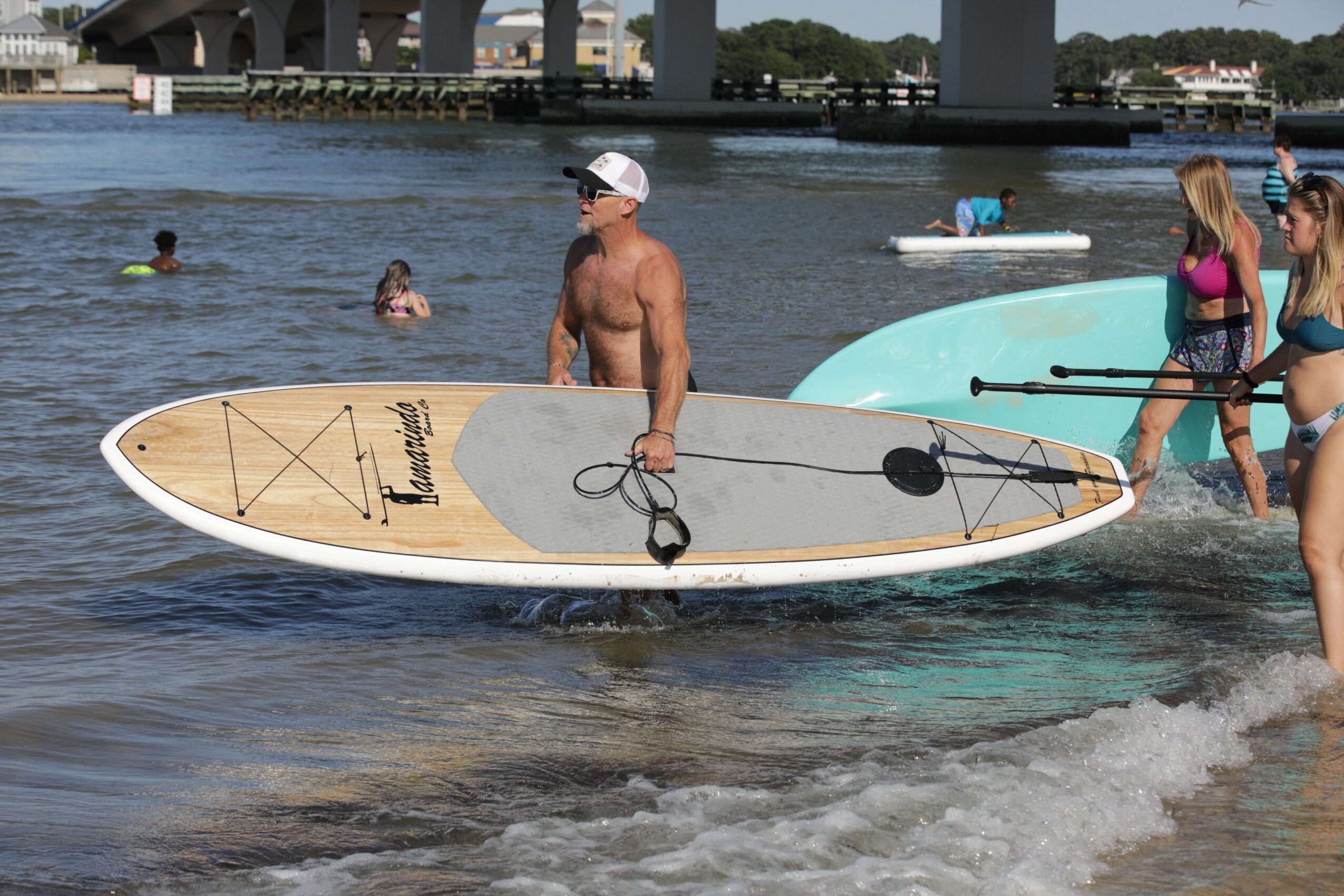 Man with a paddleboard on water, people swimming and standing nearby, bridge in background, sunny day.