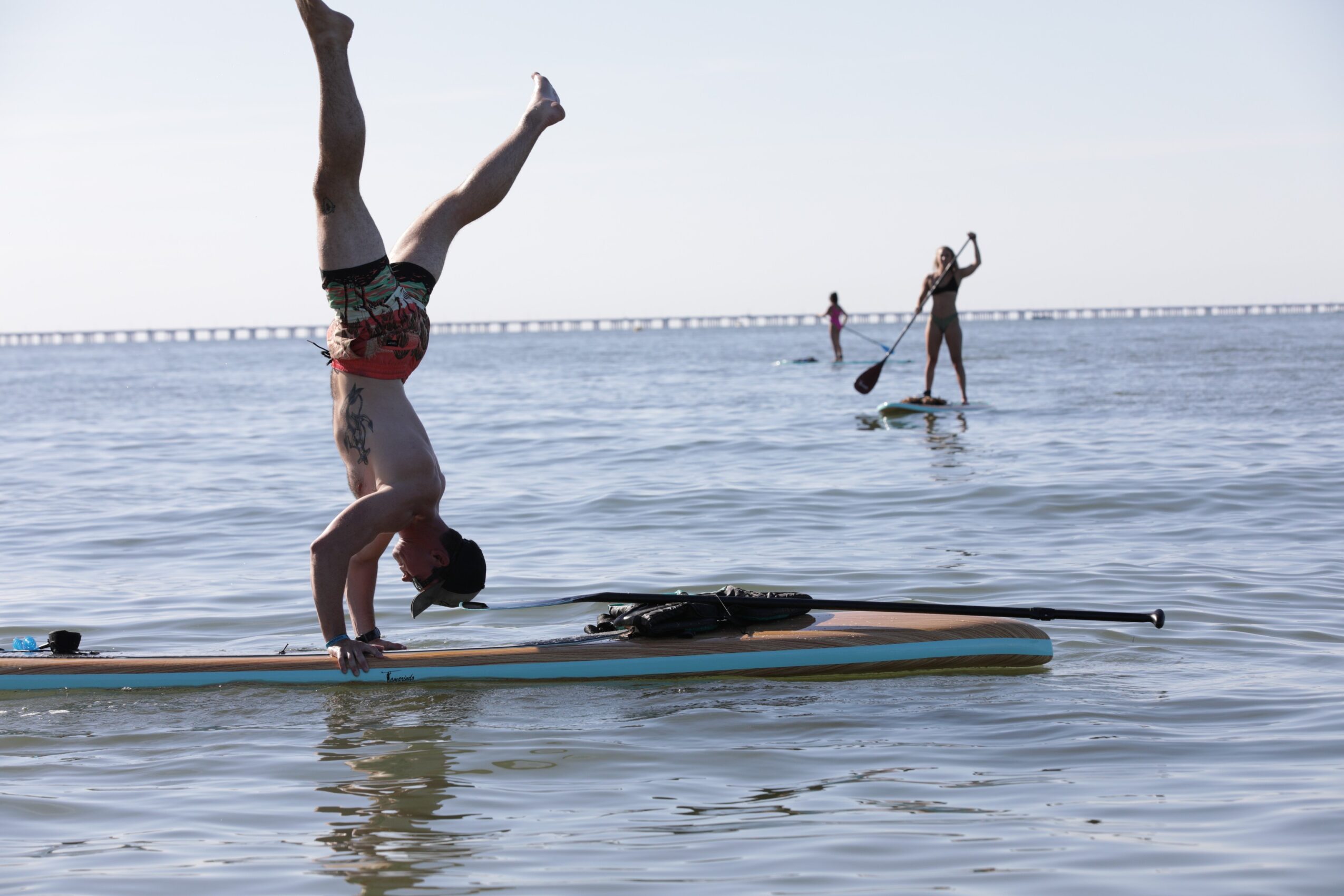 Person performing a handstand on a paddleboard in calm water, with two others paddleboarding in the background, and a bridge in the distance.