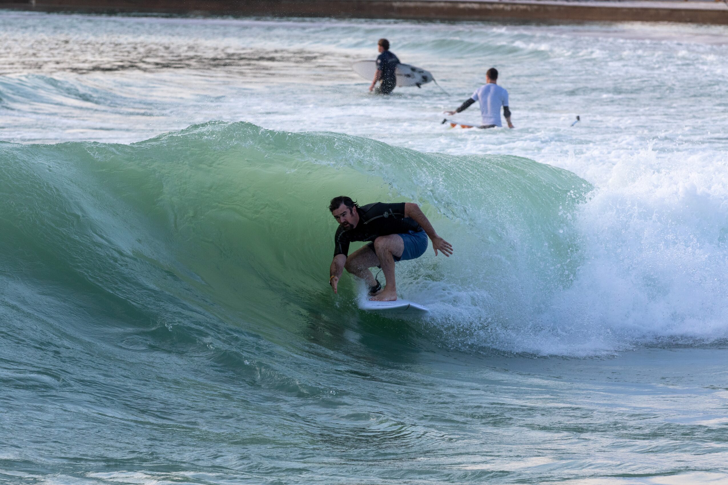 Person surfing inside a wave with two people in the background, ocean and shoreline visible.