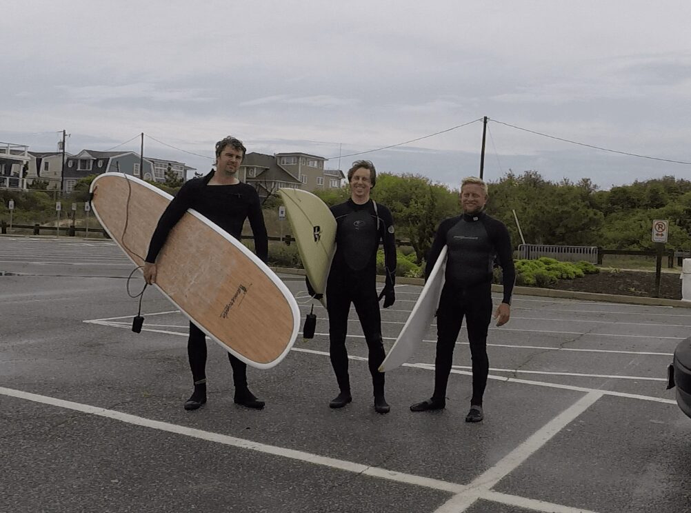 Three people standing in a parking lot holding surfboards, with cloudy sky and buildings in background.
