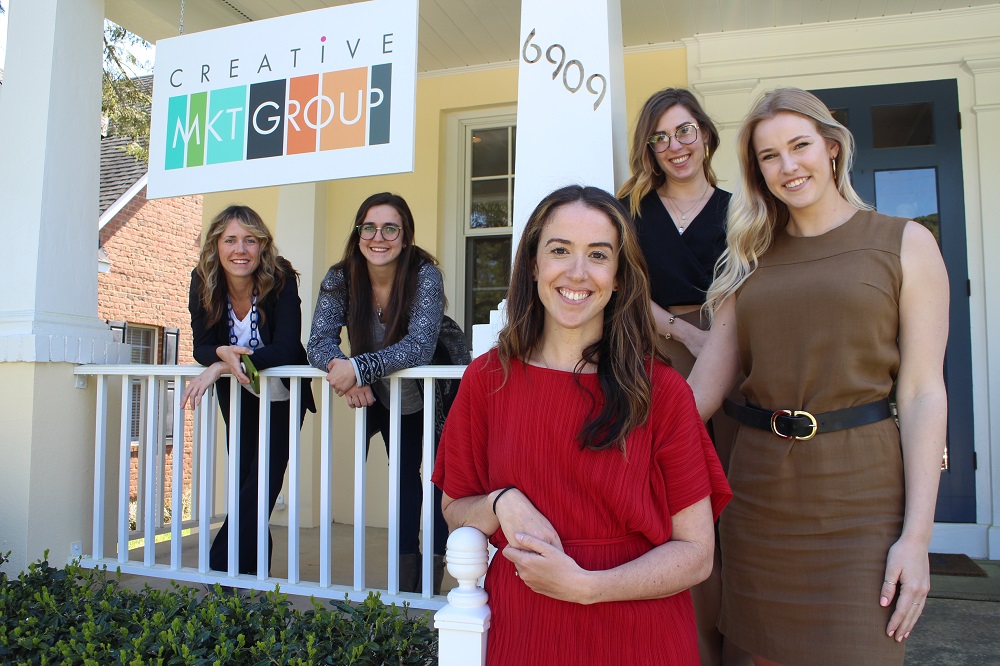 Five women standing and sitting outside a building with a sign that reads 'CREATIVE MKT GROUP.'