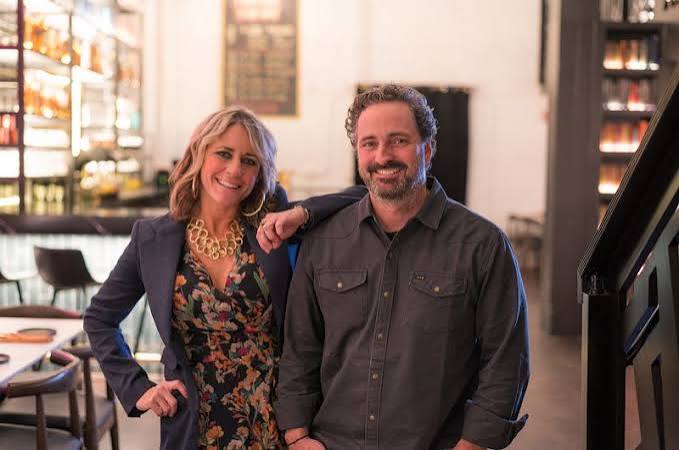 Two people, a woman and a man, smiling indoors with bookshelves in the background.