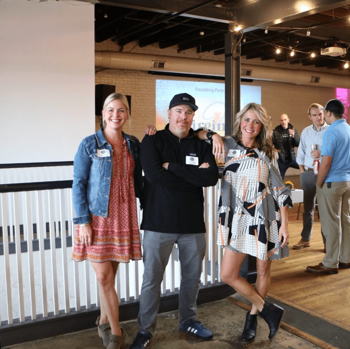 Three people standing together indoors, smiling, with a group of people in the background, railing, and ceiling lights visible.