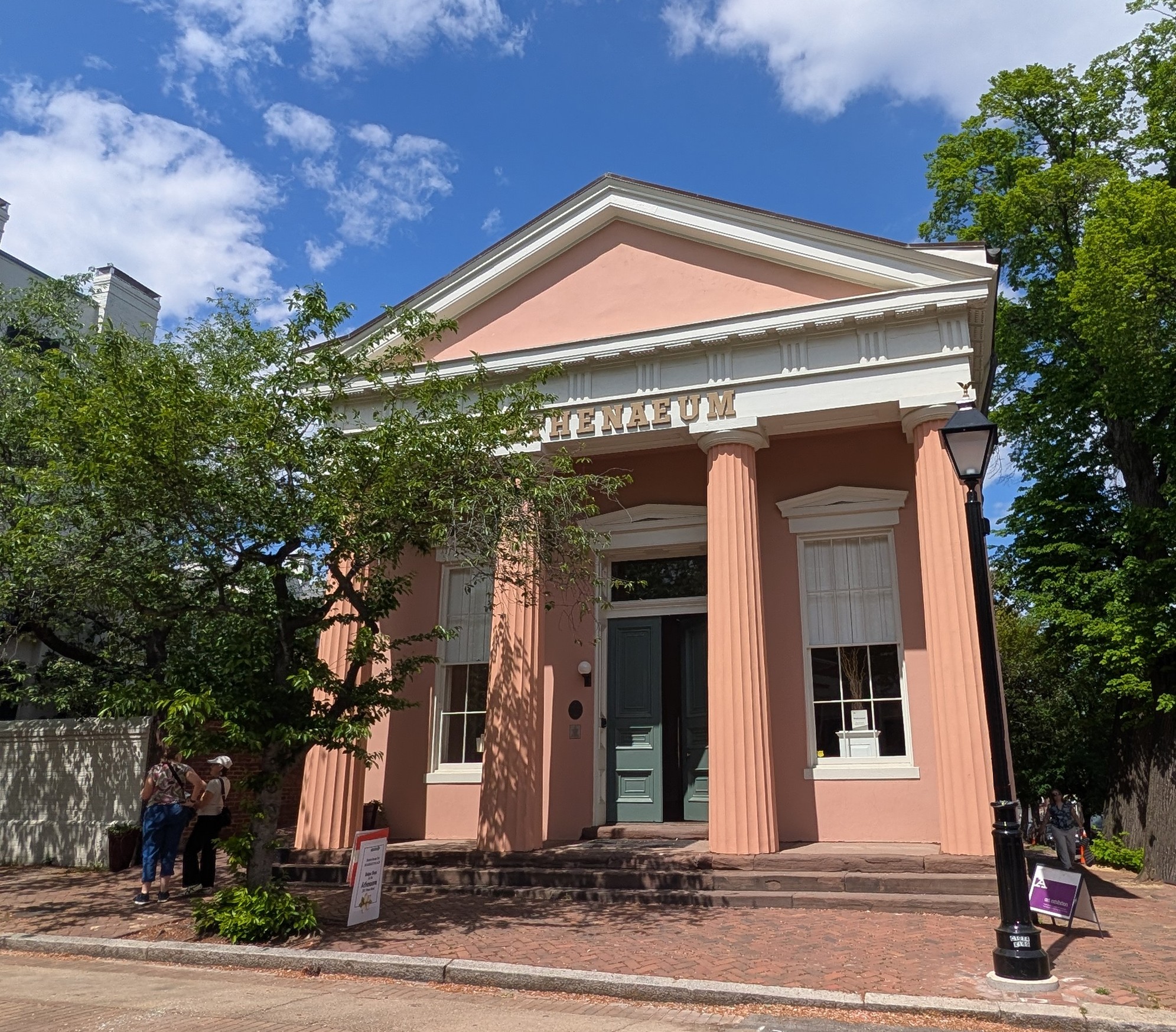 Pink building with large columns and a triangular pediment, trees and a blue sky in the background.