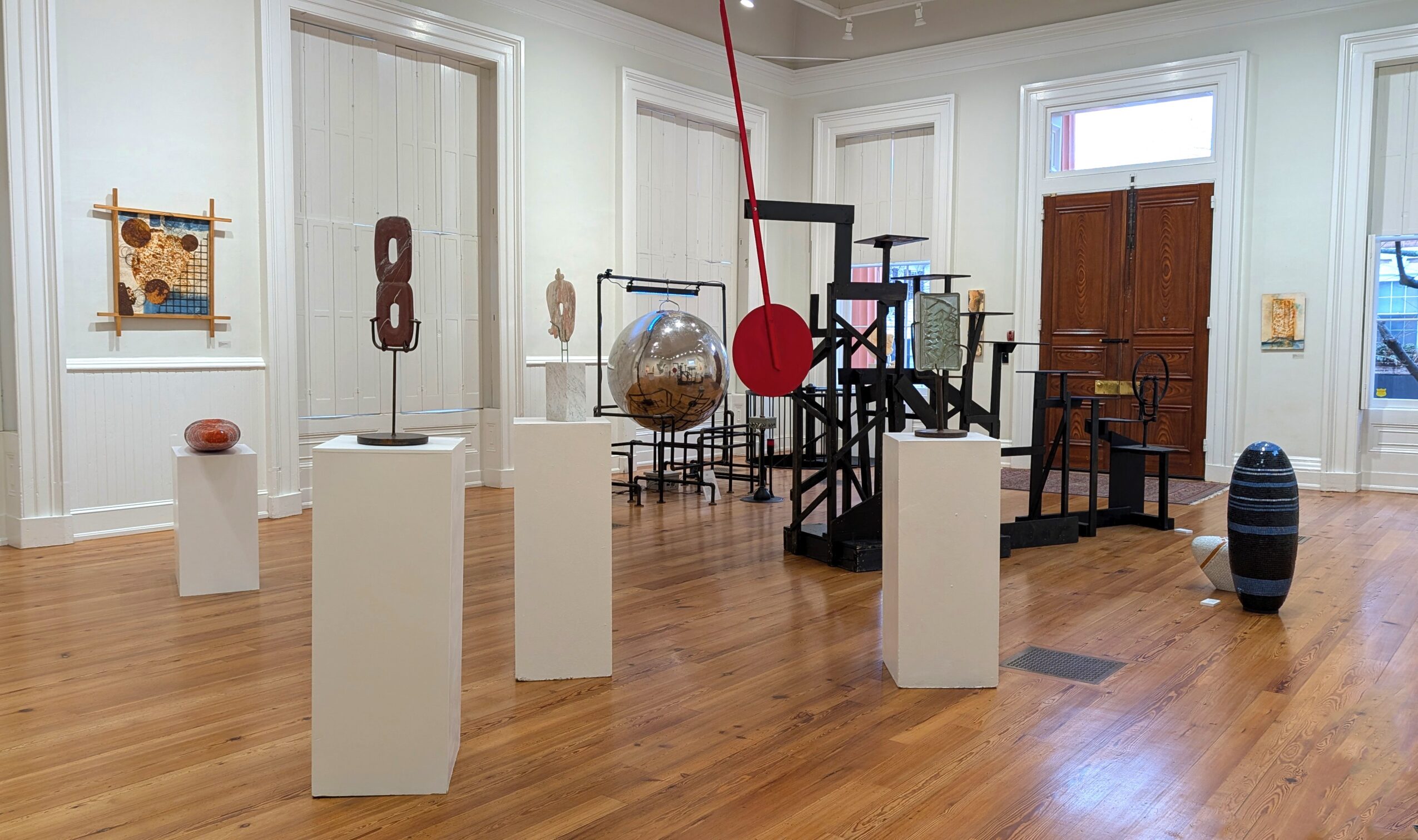 Gallery room with various sculptures and art pieces on white pedestals, wooden floor, and windows with white trim.