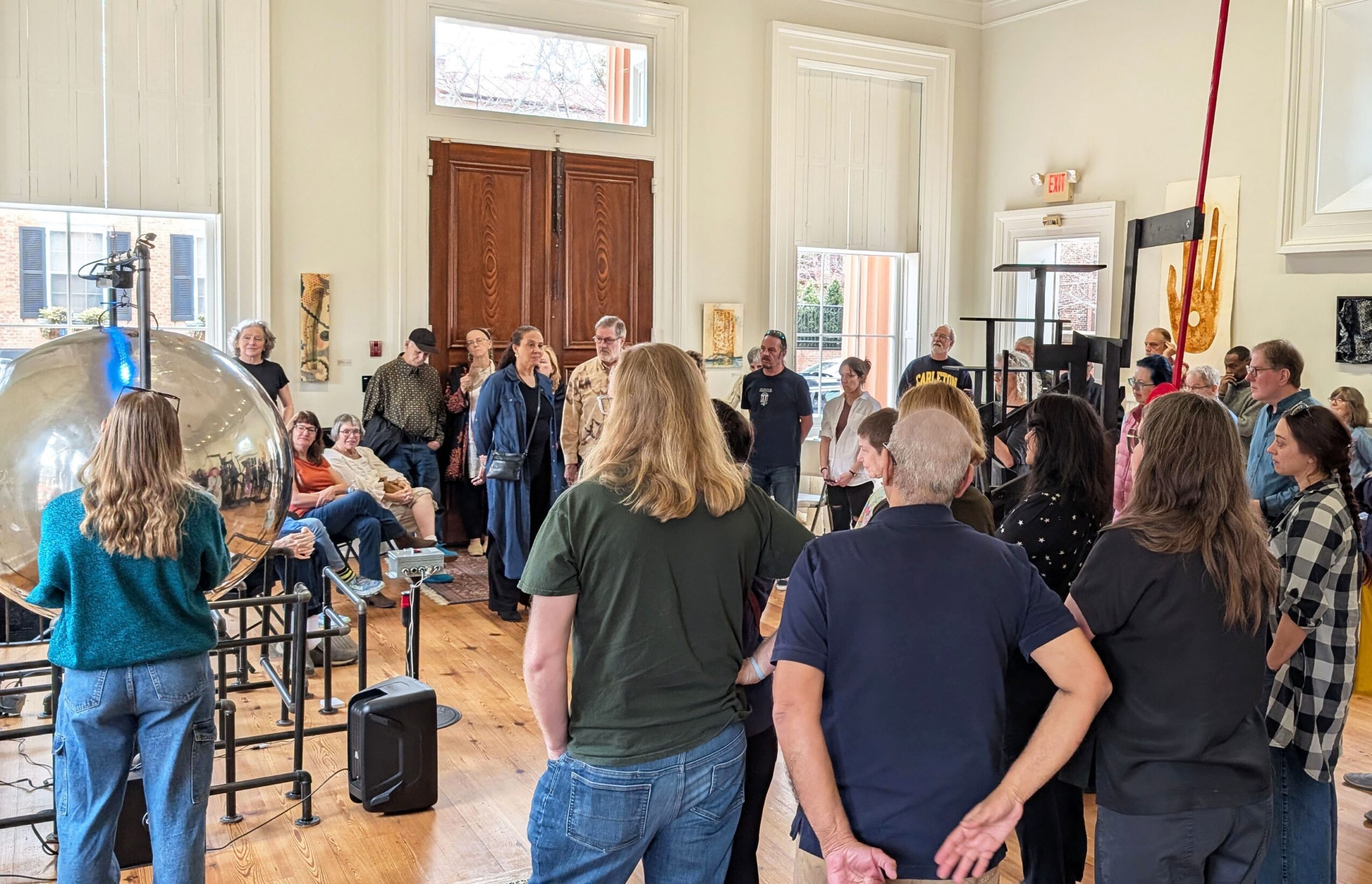 Group of people gathered in a bright room with large windows, some standing and some sitting, engaged in conversation.