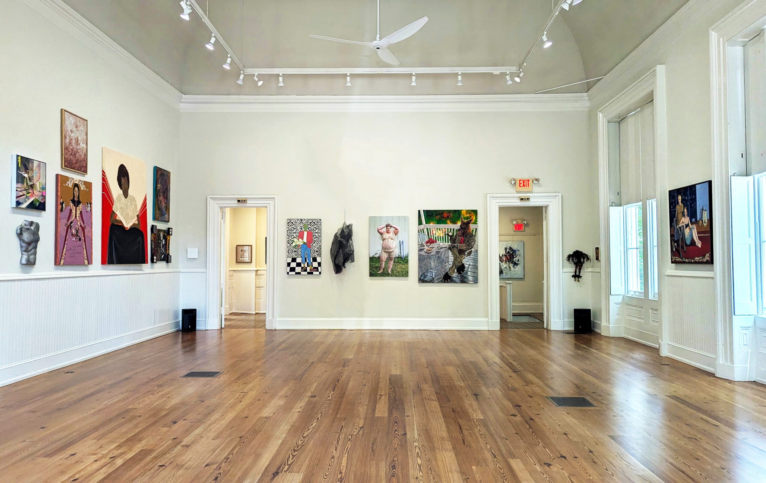 Gallery room with white walls, wooden floor, and artwork displayed on walls, with two doorways and windows.