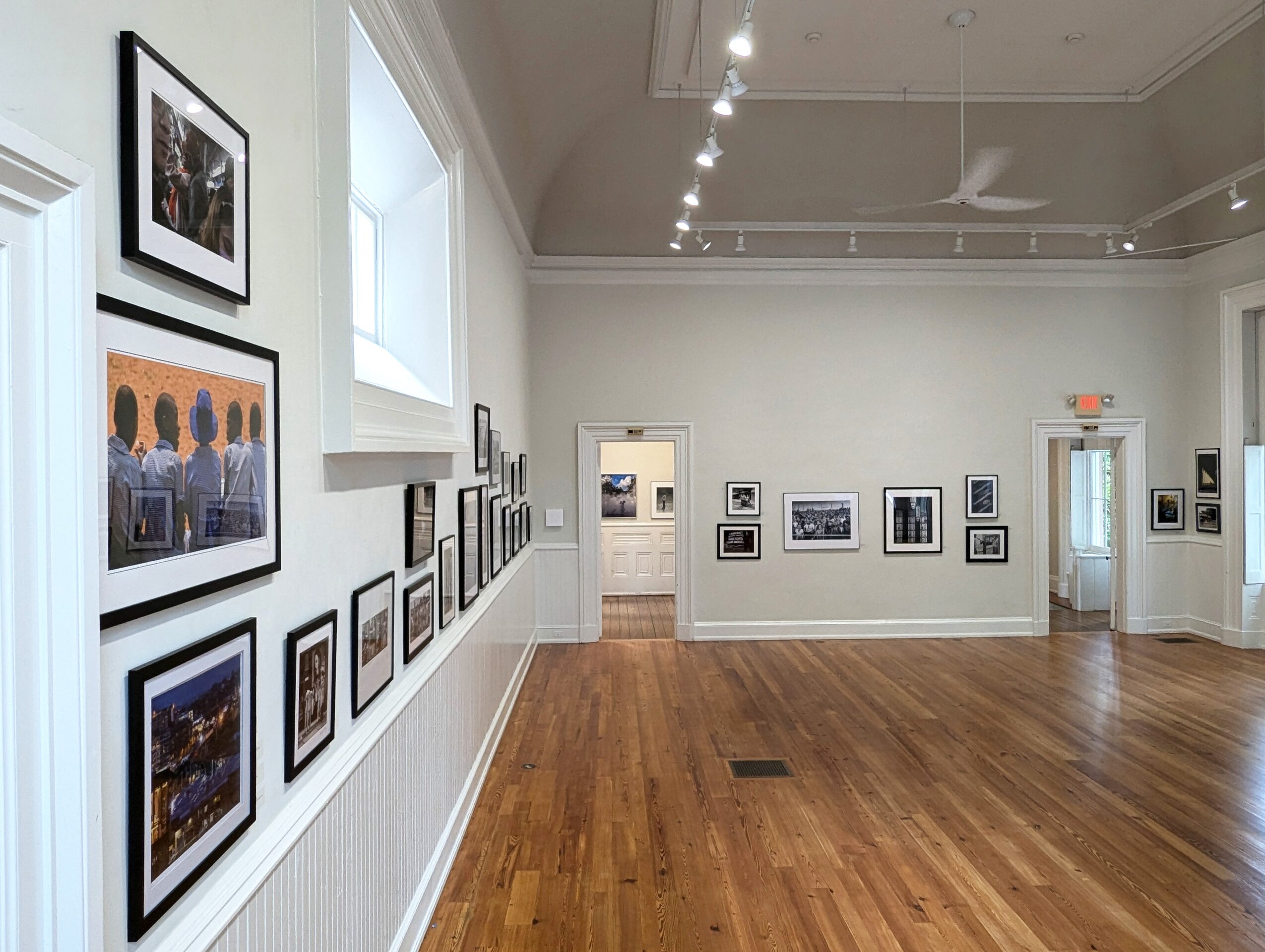 Gallery with framed artwork on white walls, hardwood floor, and ceiling lights, viewed from the left side.