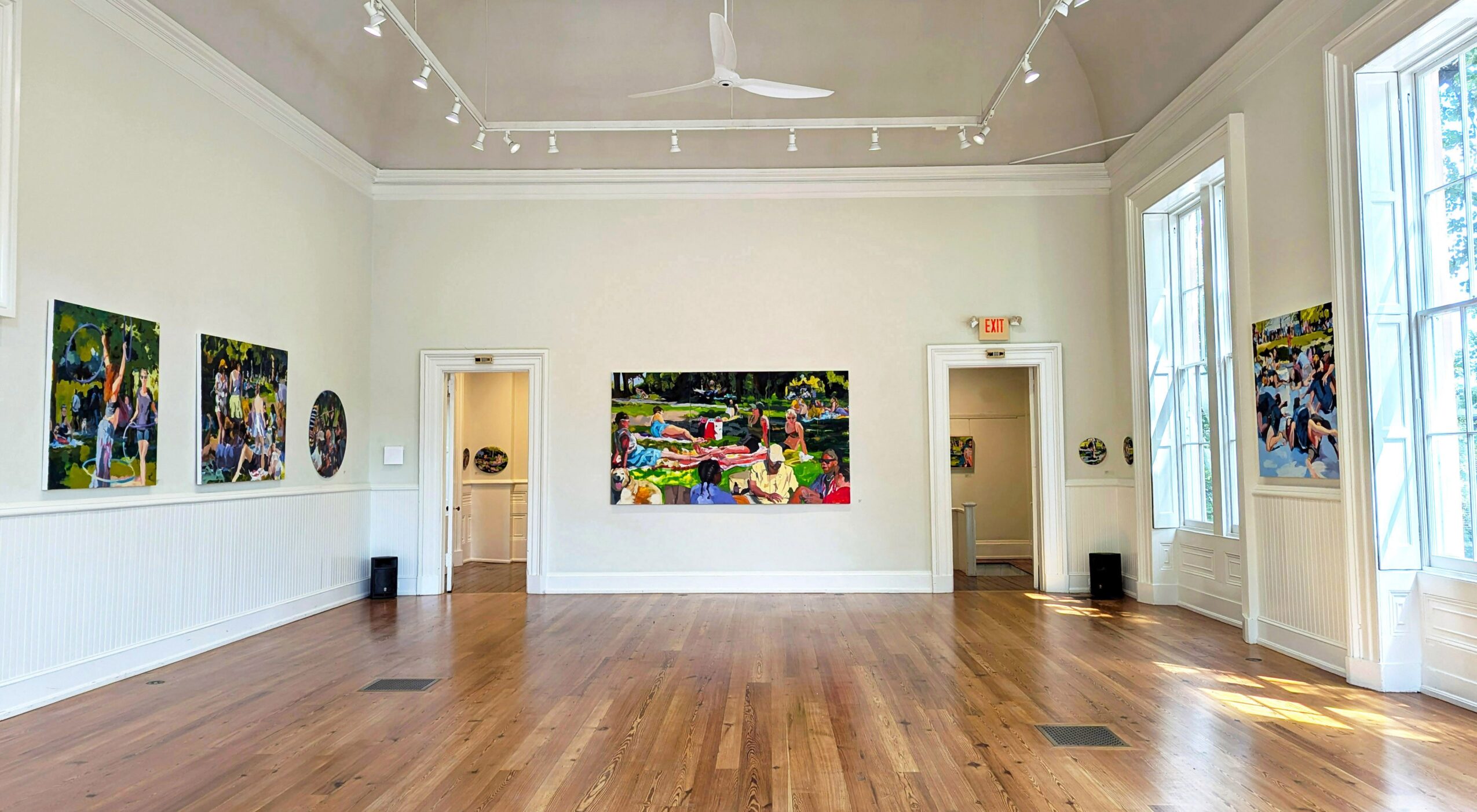 Empty art gallery with paintings on white walls, wooden floor, large windows, and high ceiling.