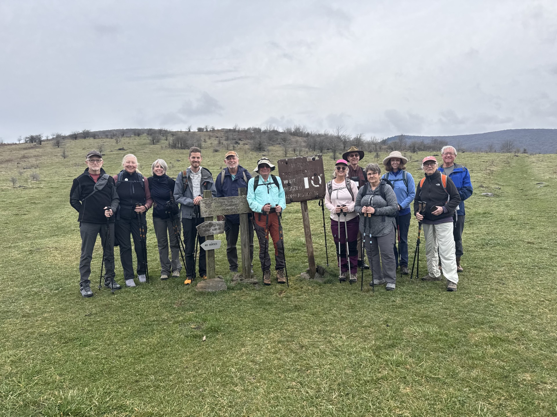 Group of ten people standing outdoors on grassy field with cloudy sky, some holding walking sticks, in a rural landscape.